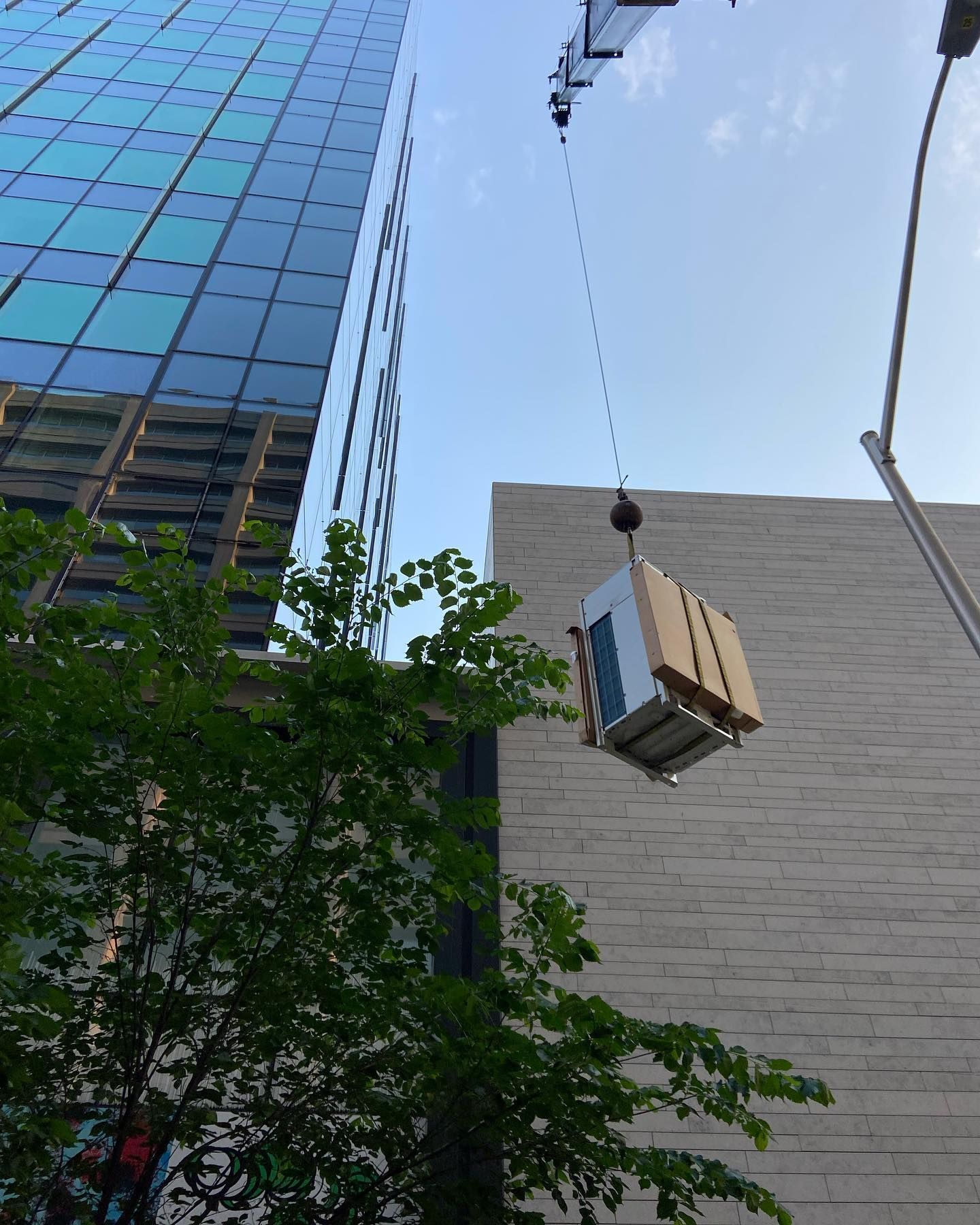 A crane hoists a large, wooden-crated object toward the top of a modern high-rise building against a bright blue sky.