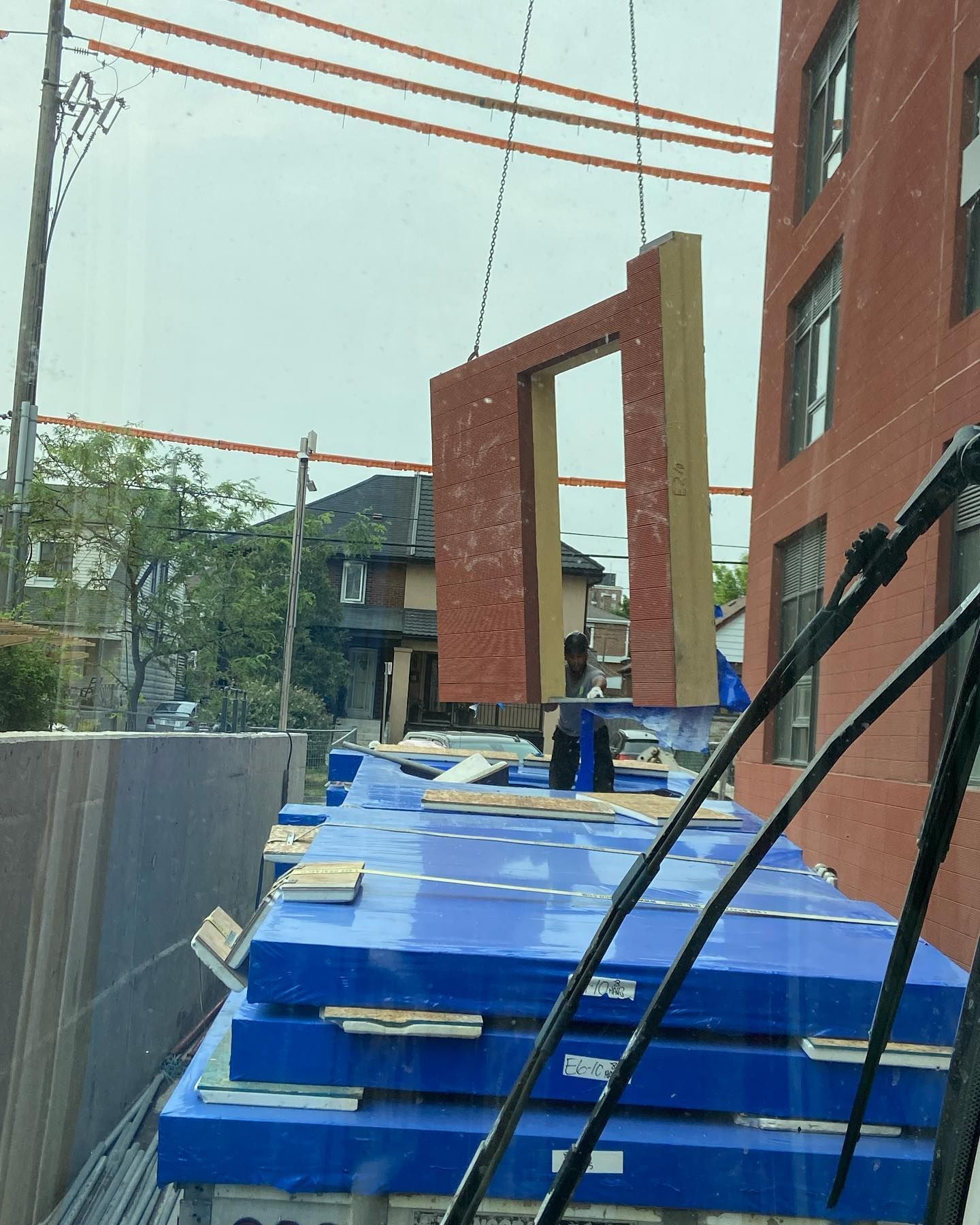 A construction crane hoists a prefabricated red brick wall panel above stacks of building materials at a job site.