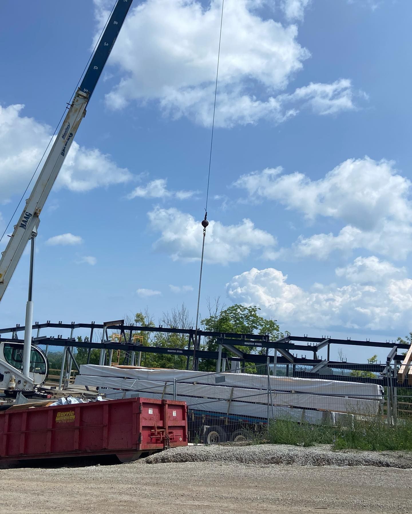 A crane lifts a stack of long white rectangular panels at a construction site under a partly cloudy blue sky.