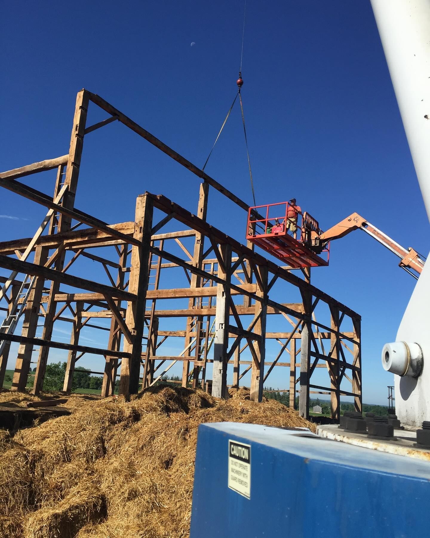 A crane lifts a wooden frame component above a construction site, with a worker in a boom lift assisting the process.