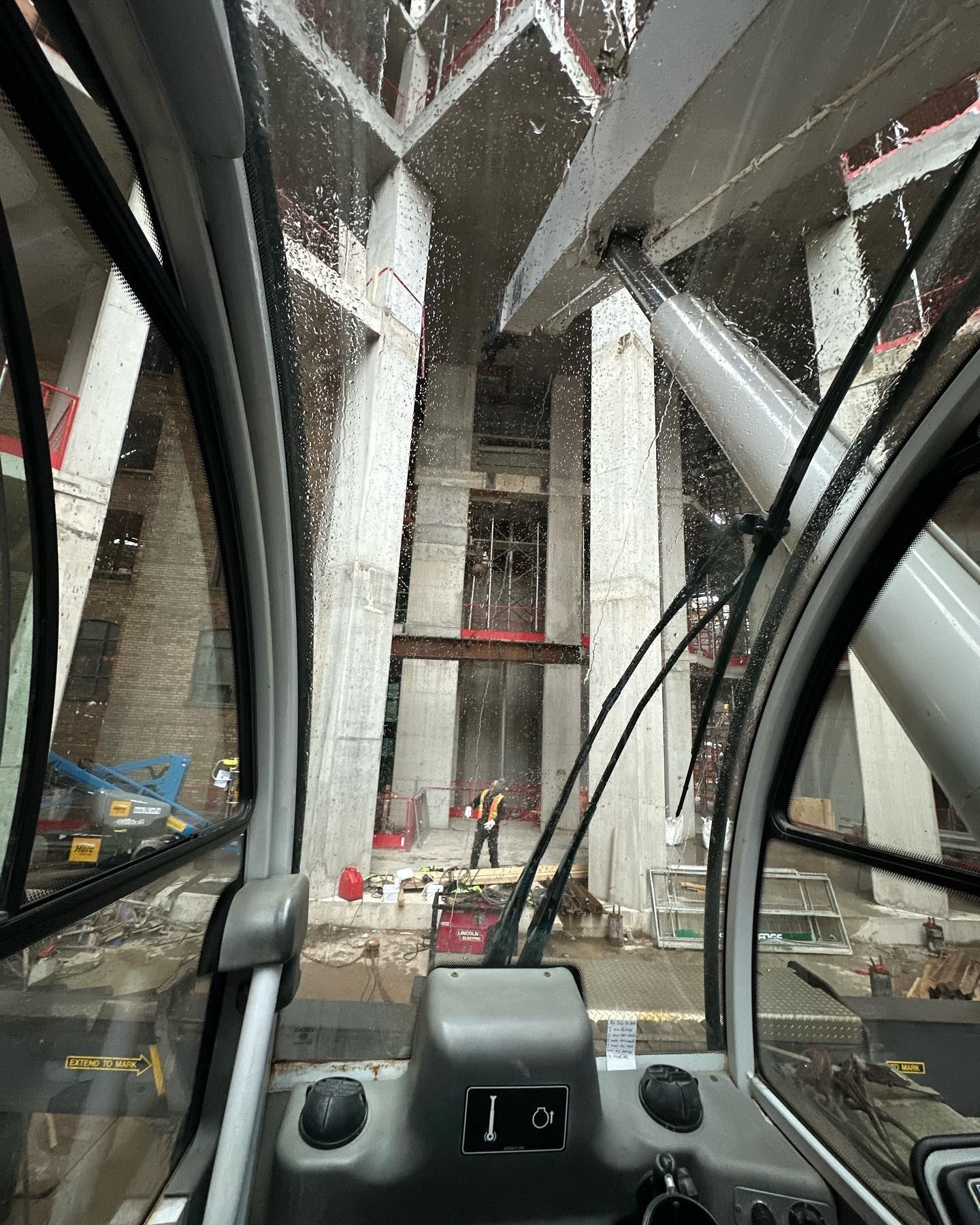 View from a construction vehicle cab showing a worker standing in a concrete structure under construction.