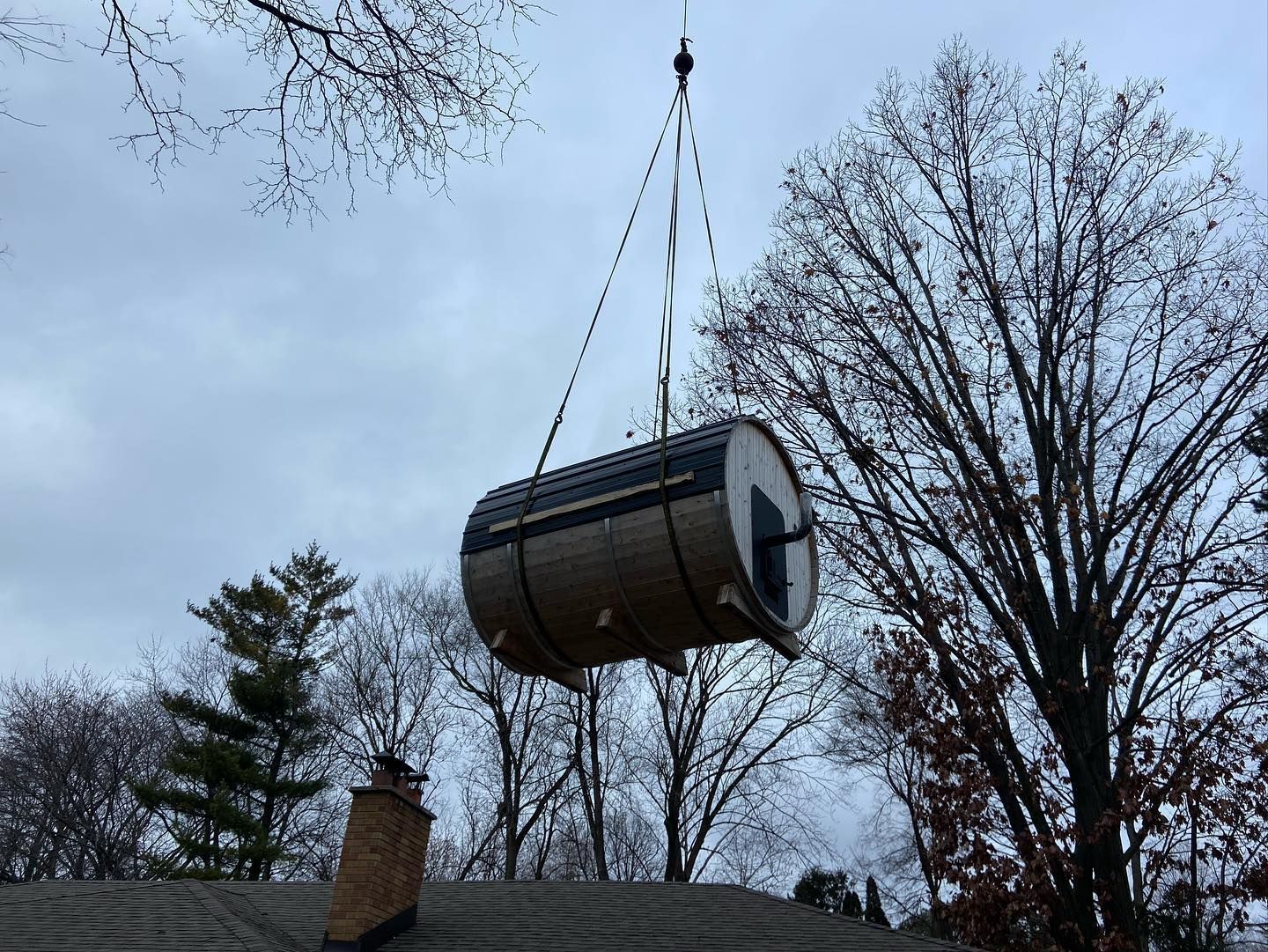 A crane lifts a cylindrical wooden barrel sauna through the air above a residential building against a cloudy sky.