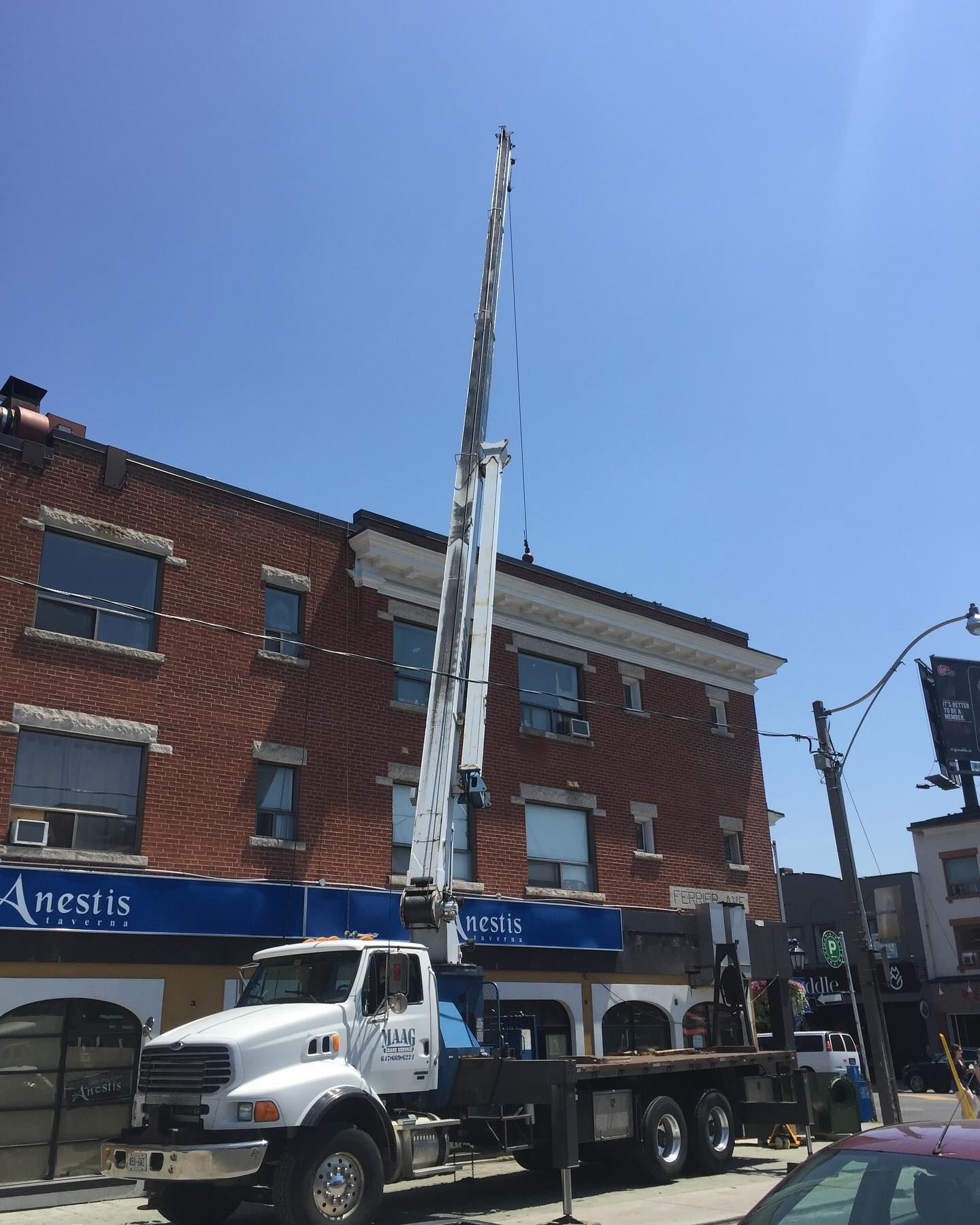 A white utility truck with an extended boom crane parked on a street in front of a three-story red brick building.