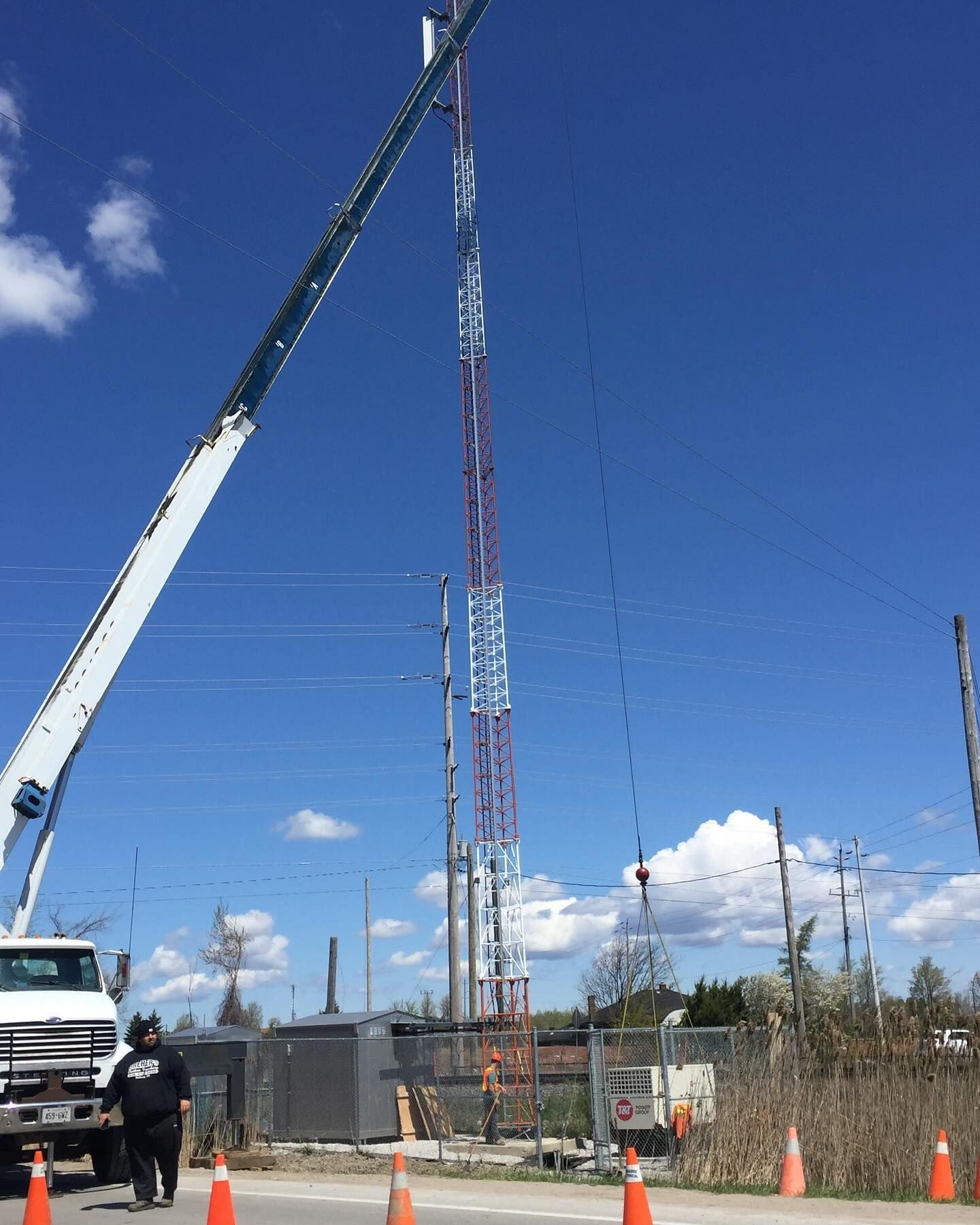 A construction crane lifts a tall communications tower against a blue sky, with orange traffic cones in the foreground.