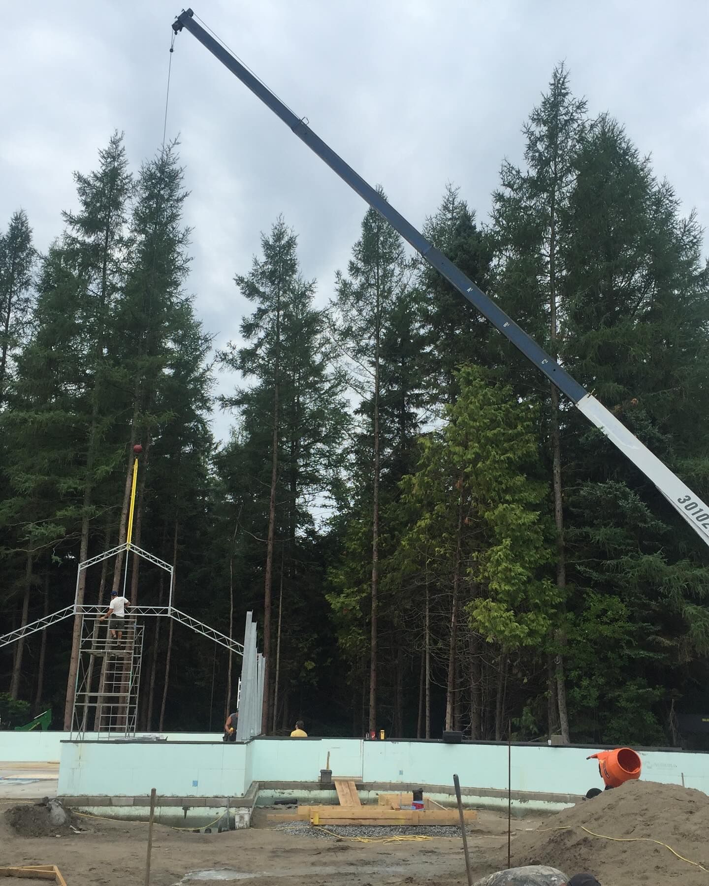 A crane lifts a structural frame over a concrete foundation on a construction site surrounded by tall pine trees.