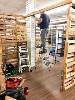 Person standing on a step stool, working inside a wooden pallet-built structure in a workshop.