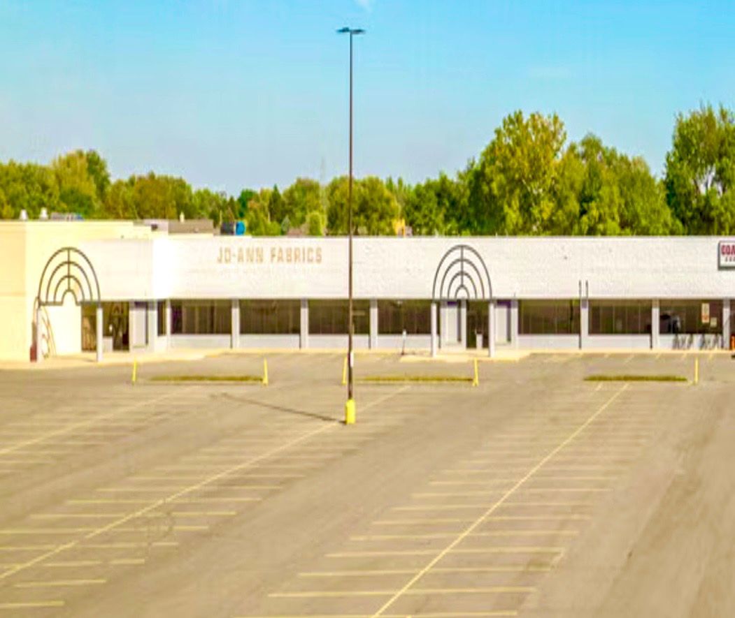 Empty parking lot in front of a white storefront with arched entrances and trees behind it.