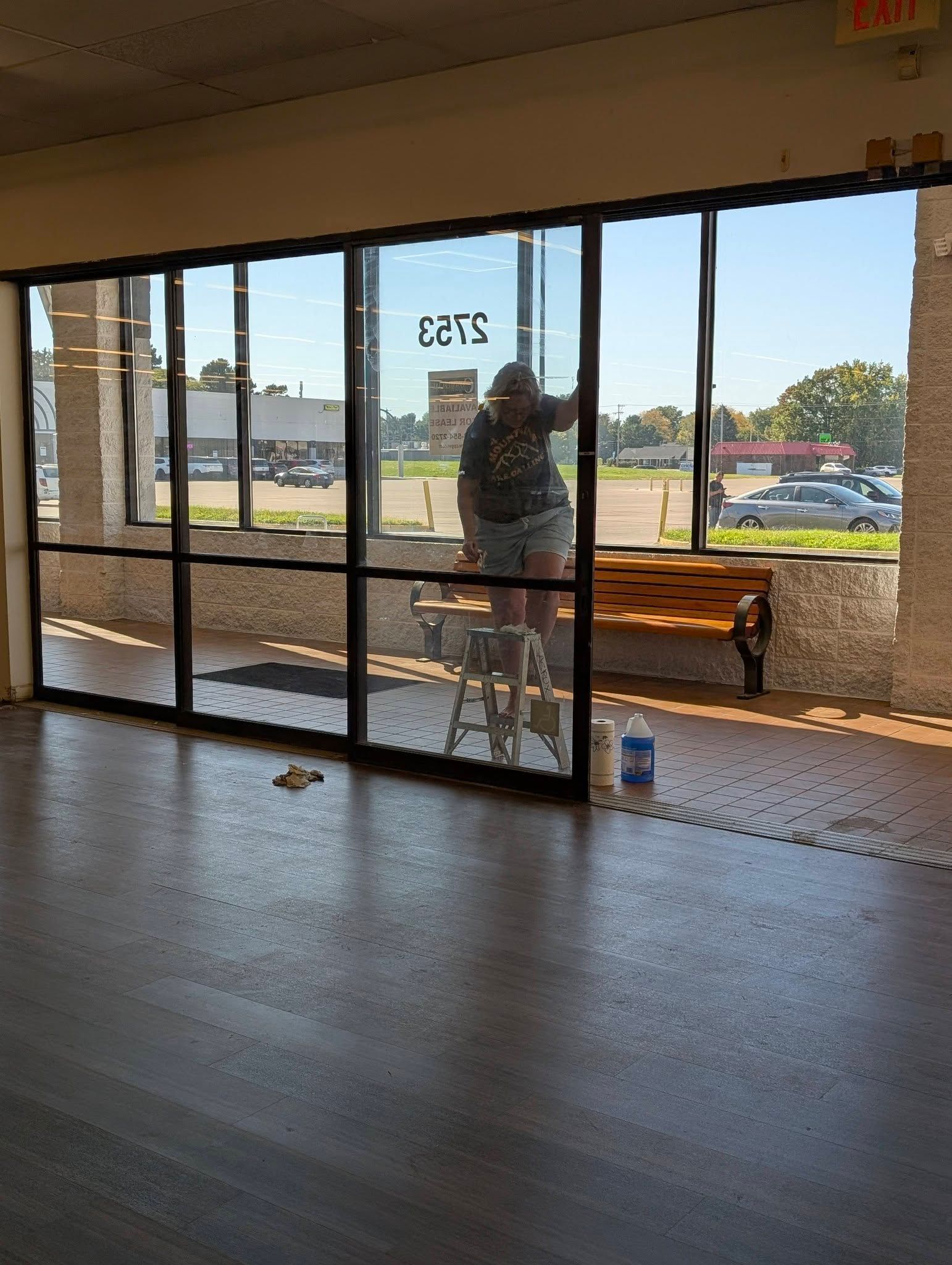 Empty storefront interior with glass doors, a bench outside, and a person standing by the entrance.