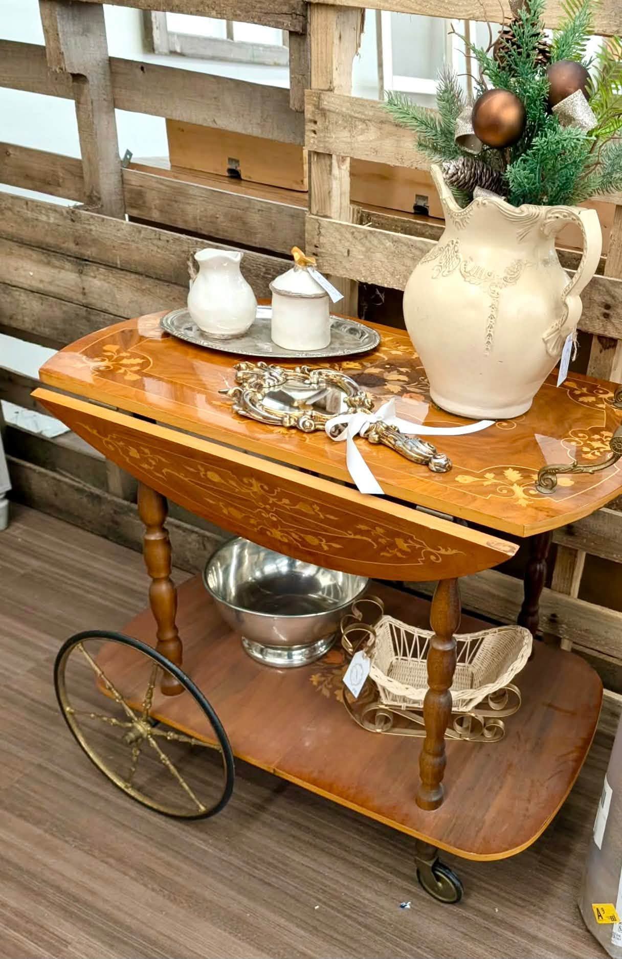 Vintage tea cart with white teapot, cups, and a small floral arrangement against a rustic wood fence