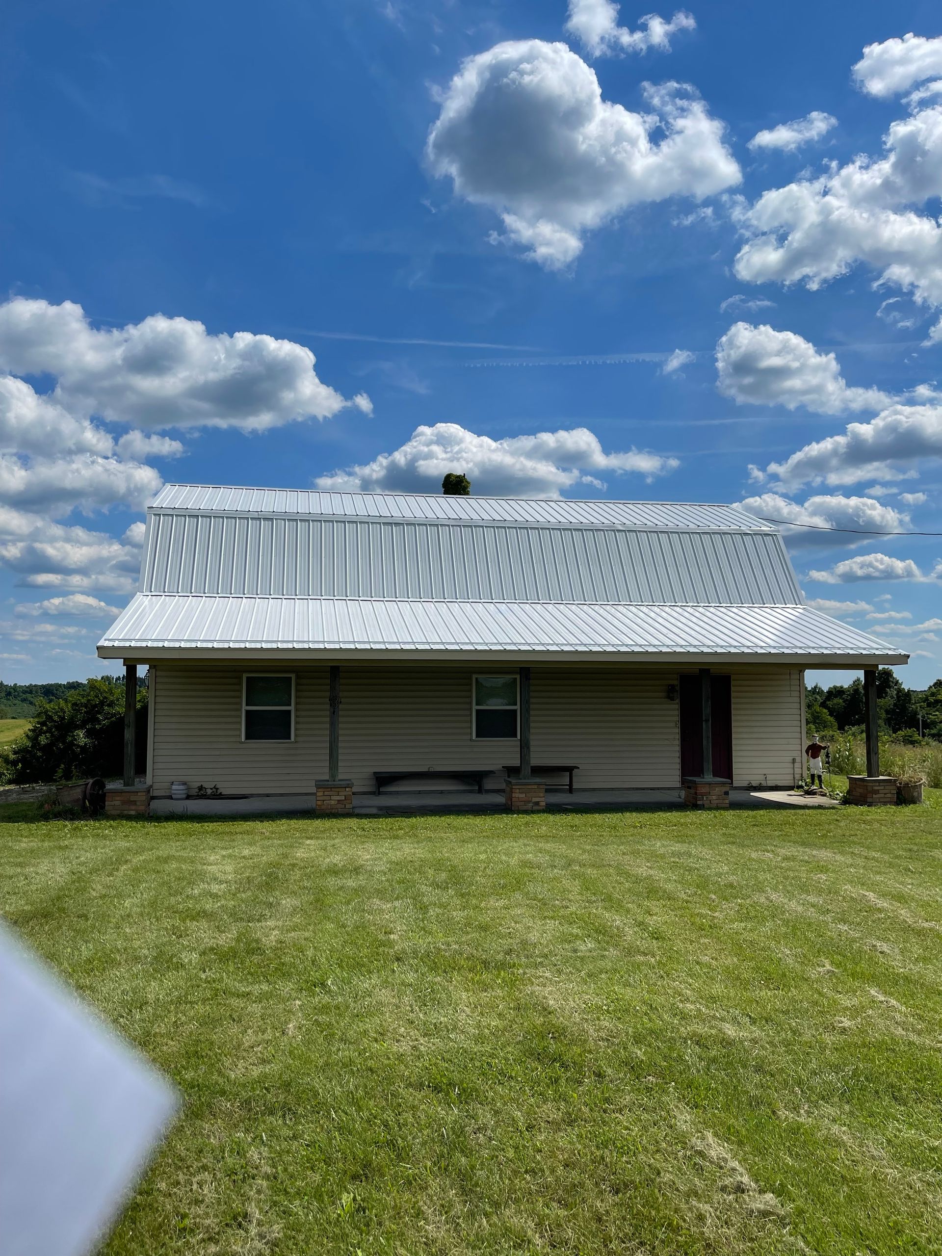 Small rural house with a metal roof under a bright blue sky with clouds, viewed over a grassy yard.