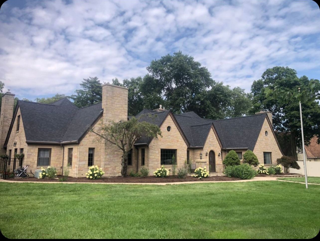 Stone suburban house with dark roofs, central chimney, and a green lawn under a cloudy sky
