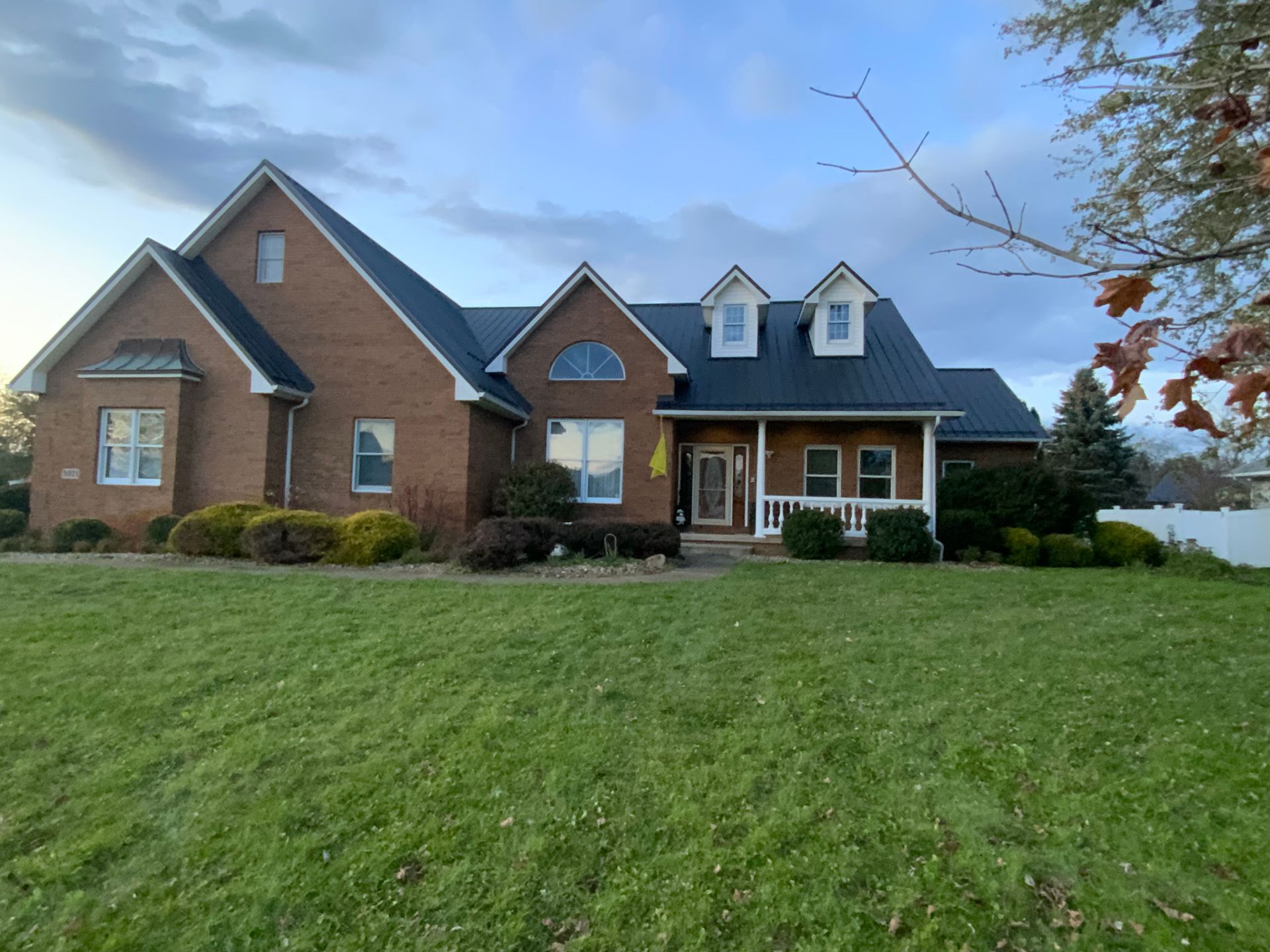 Large brick suburban house with a dark roof, front porch, and a wide green lawn under a blue sky