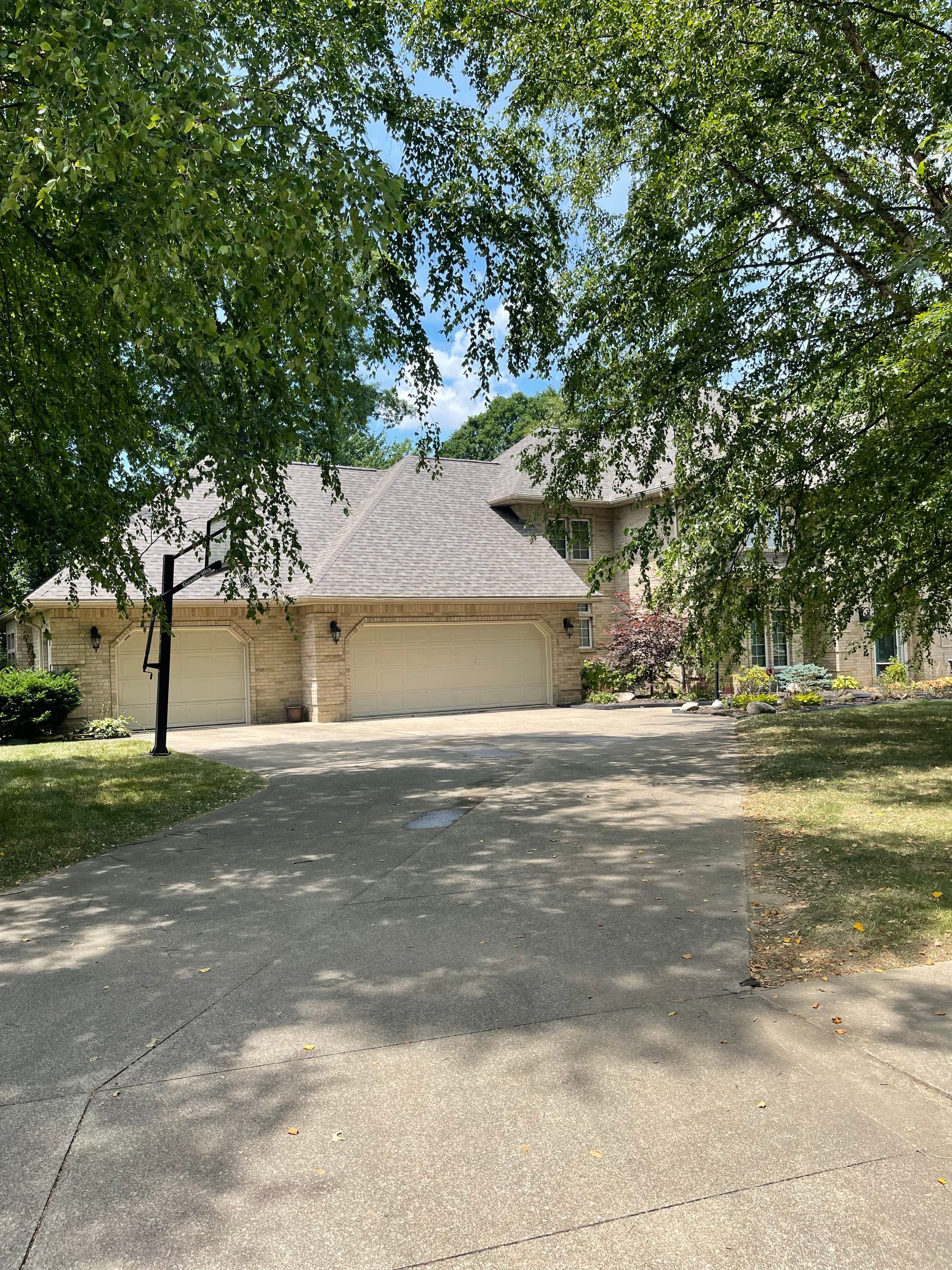 Gravel driveway leading to a beige house with a two-car garage, framed by leafy trees on a sunny day
