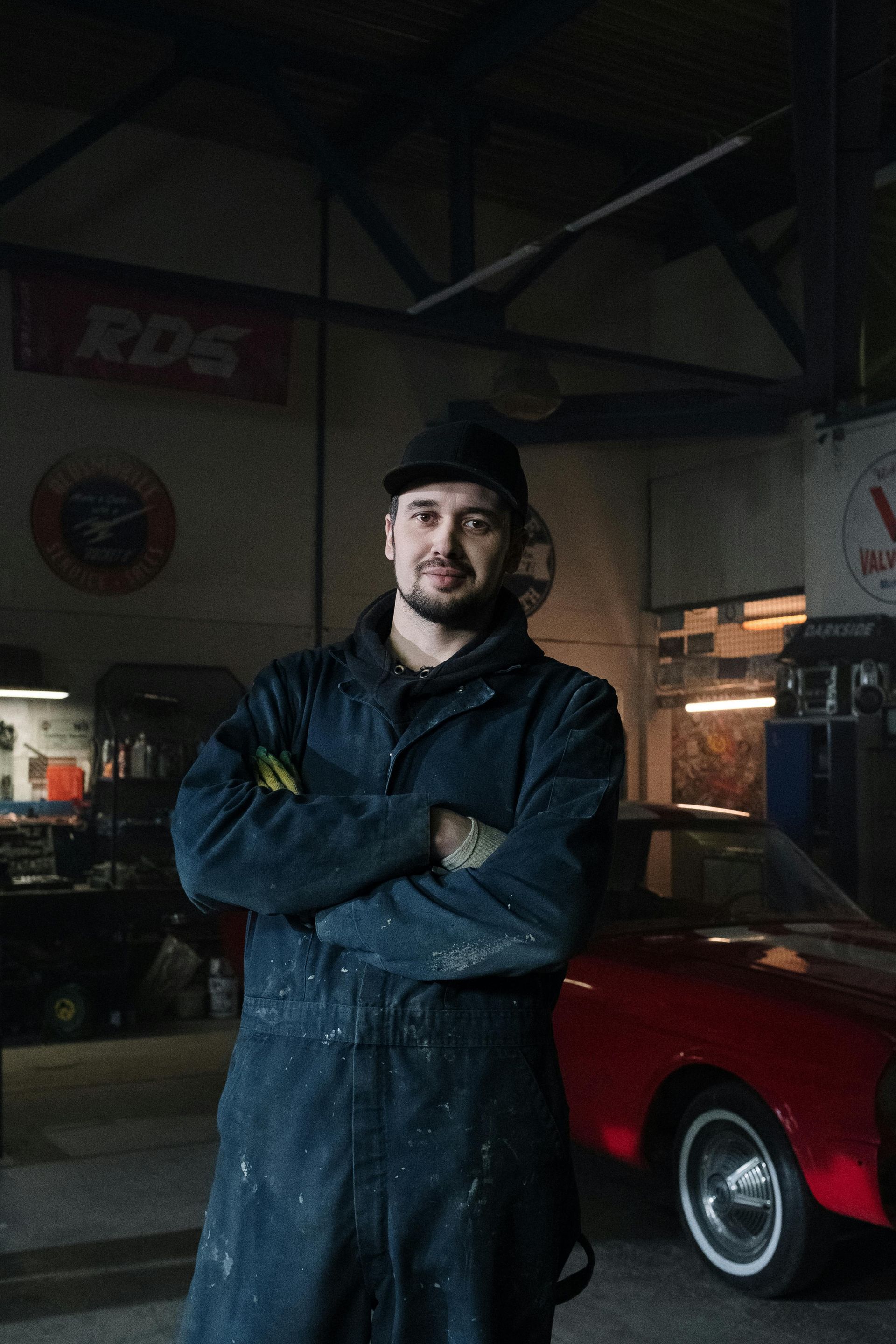 Mechanic in blue overalls, arms crossed, stands in a garage beside a red car.