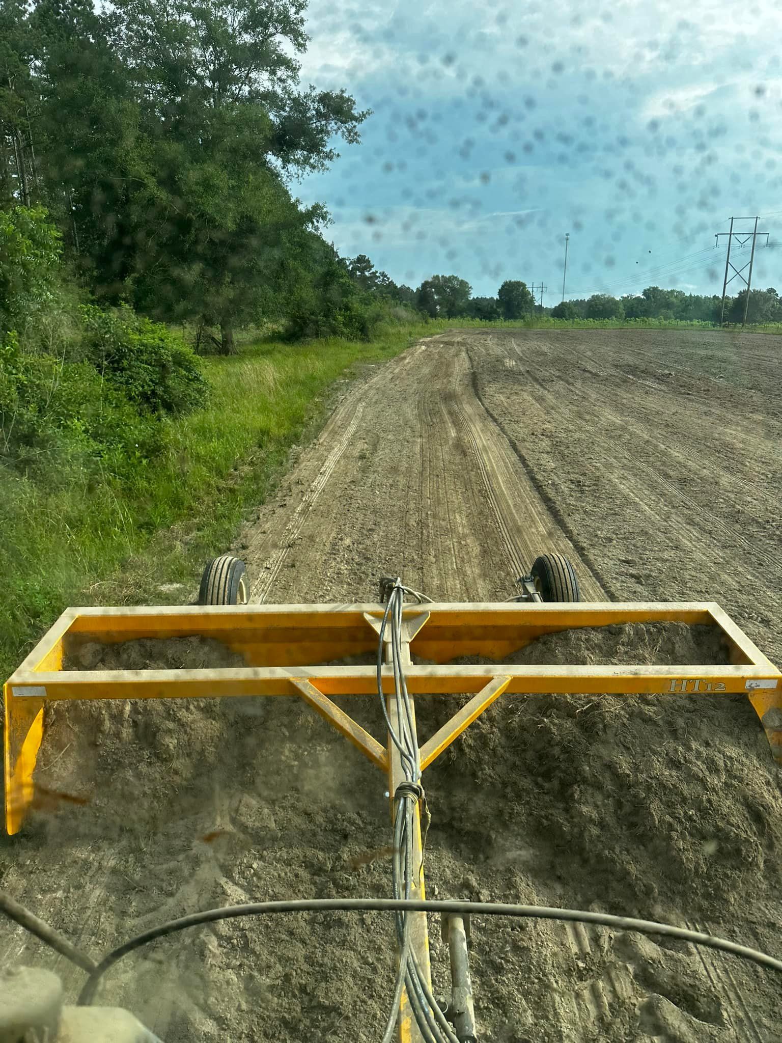 Yellow grader leveling a freshly plowed field with trees in the background.