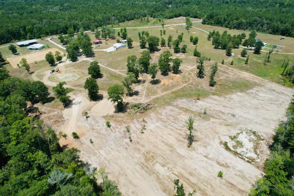 Aerial view of a cleared field with scattered trees, buildings, and surrounding forest.