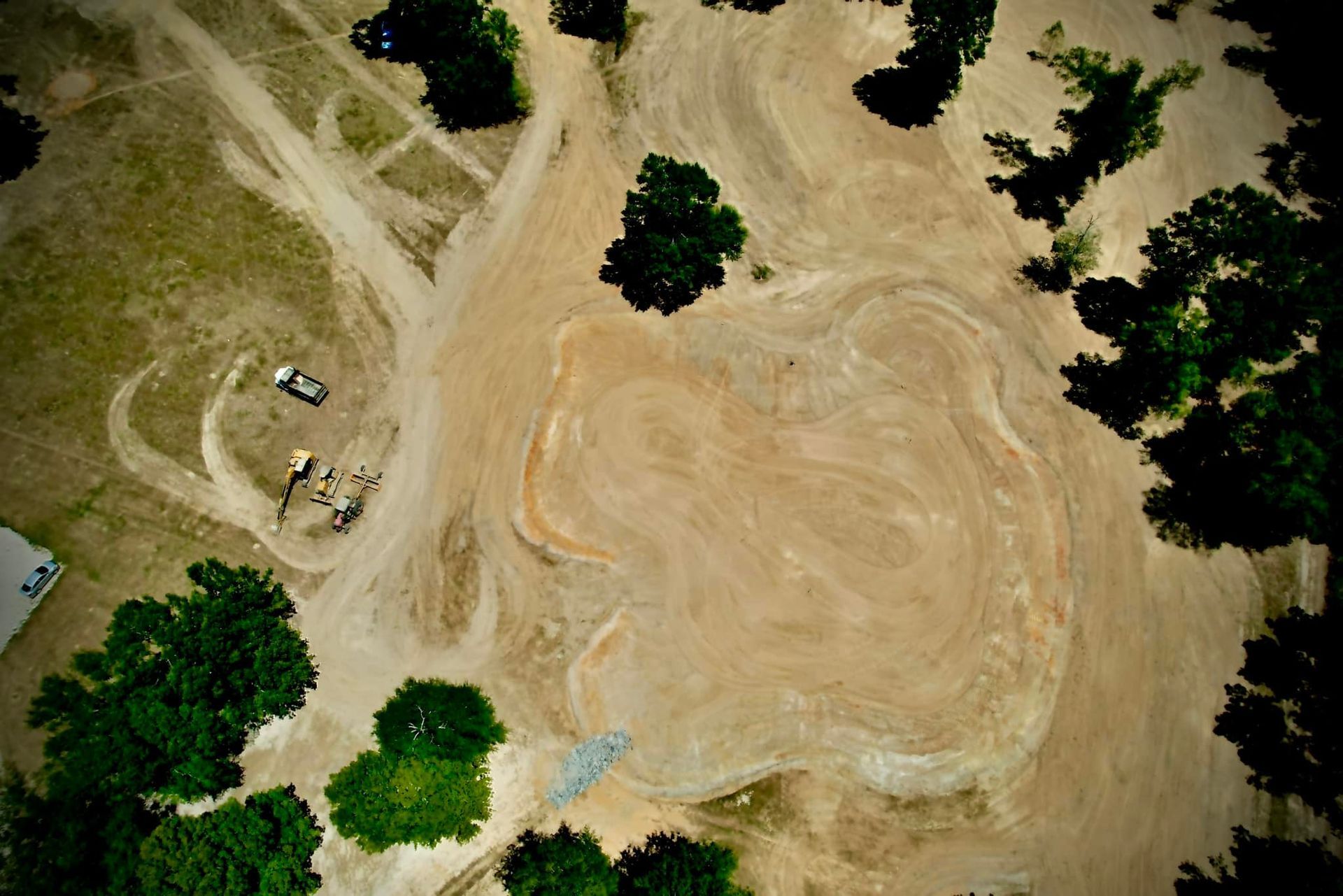 Aerial view of a dirt clearing with tree cover, construction equipment, and tire tracks.