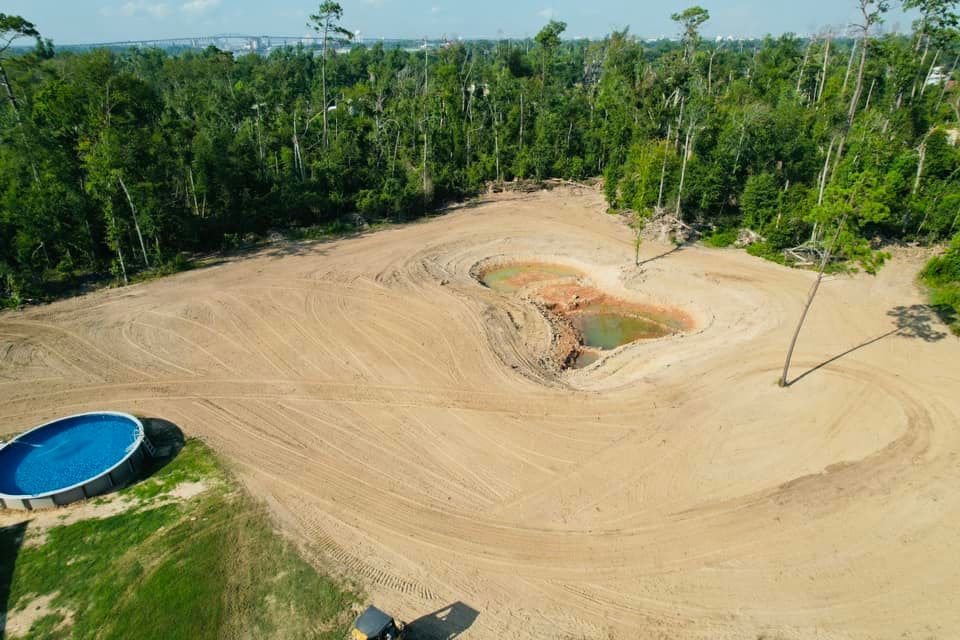 Cleared land with dirt mounds and a small body of water, with an above-ground pool and forest backdrop.