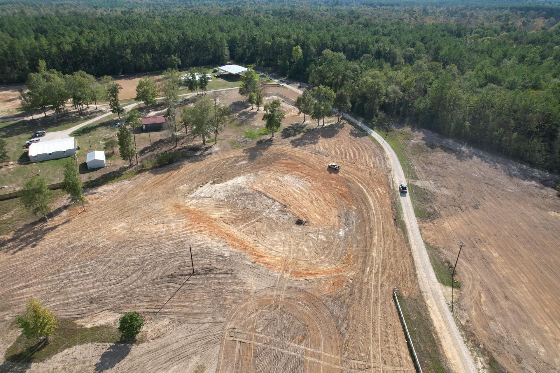 Aerial view of a cleared, brown field with a road. Several buildings and a forest border the field.