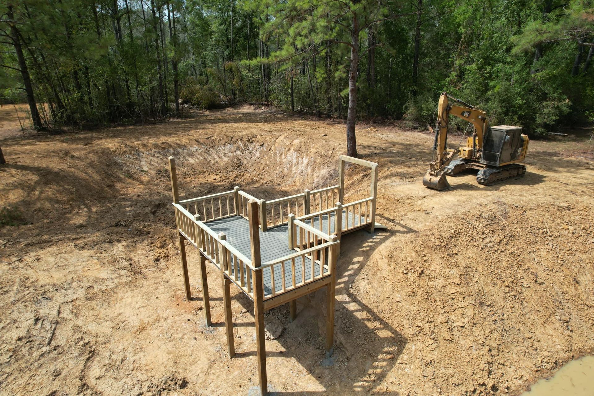 Wooden deck structure near a muddy pond, excavator in the background, forest setting.