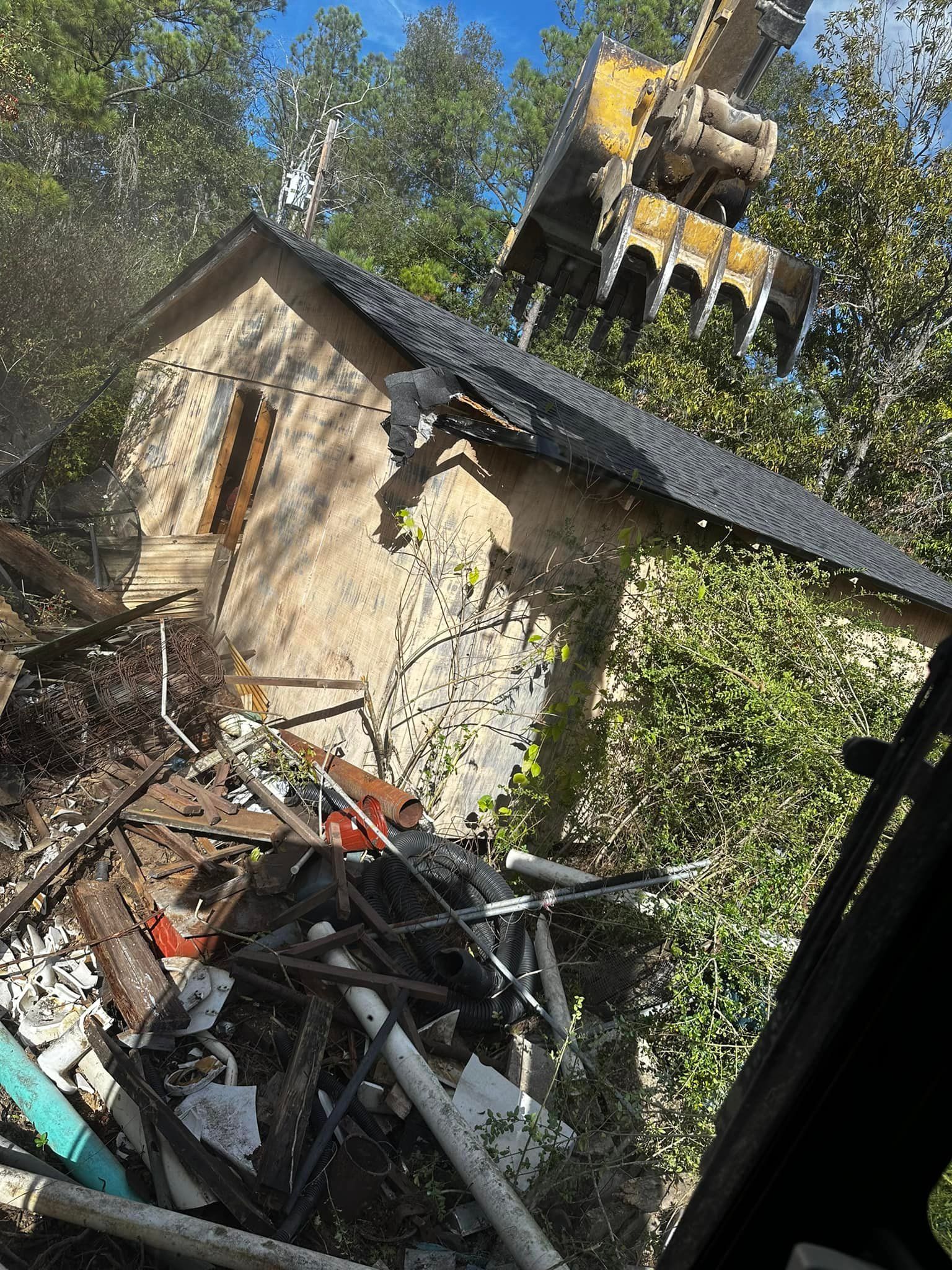 Excavator demolishing a small, tan-colored building with a dark roof, surrounded by debris and trees.