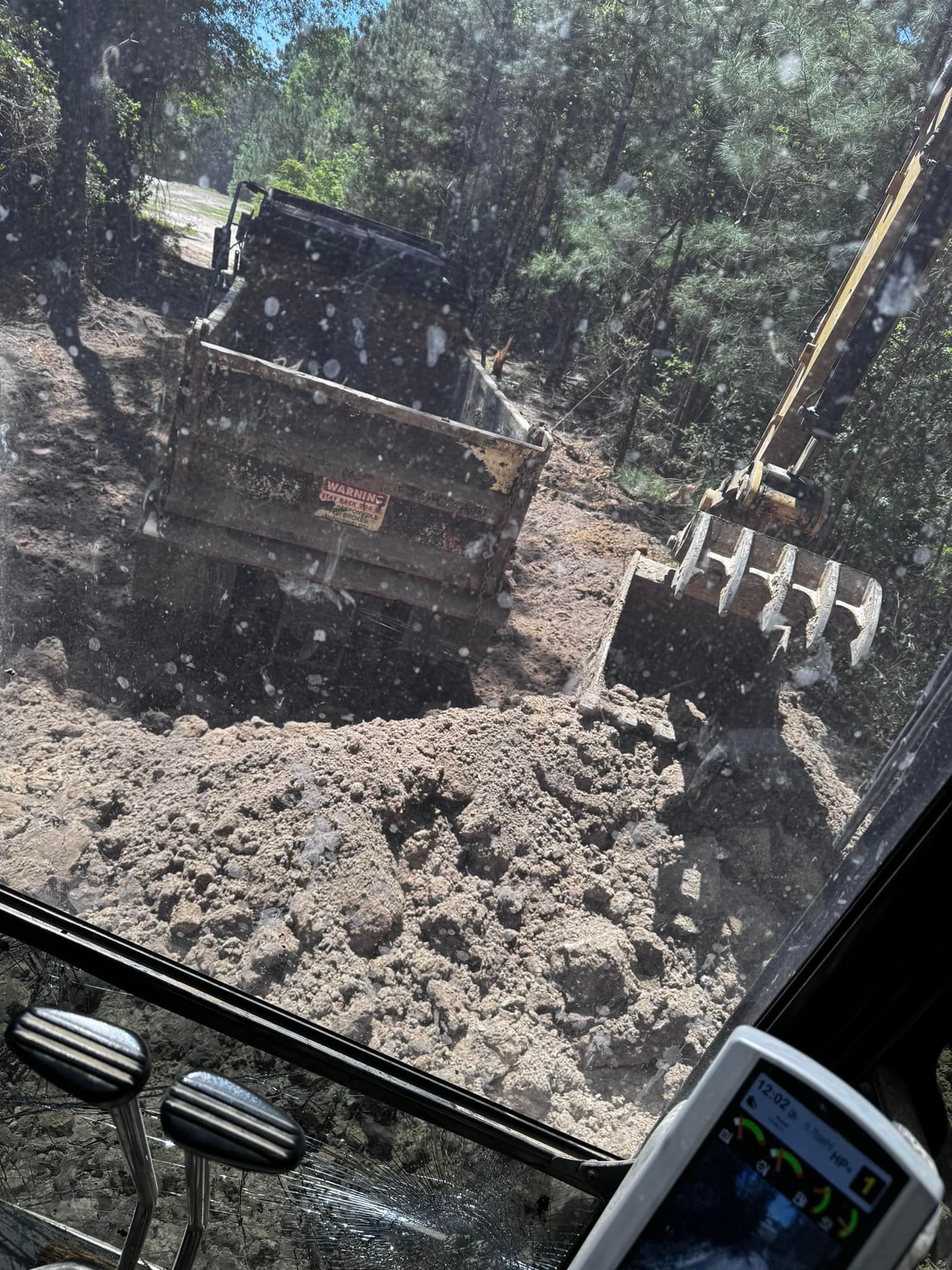 Excavator dumping soil into a truck, seen from inside the cab. Muddy brown ground, forest background.