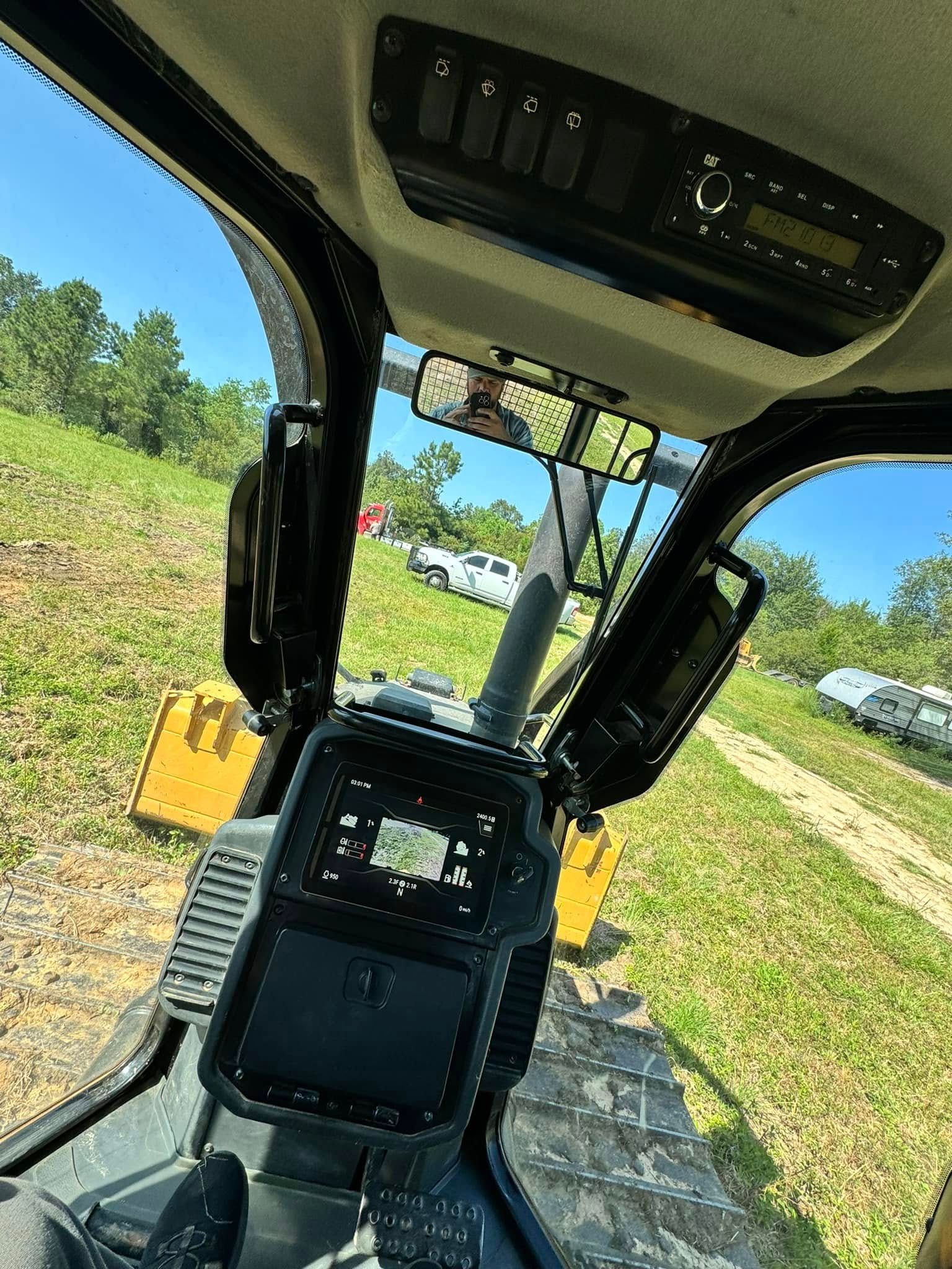 Interior view of a heavy machinery cab overlooking a grassy field with trees and vehicles in the distance.