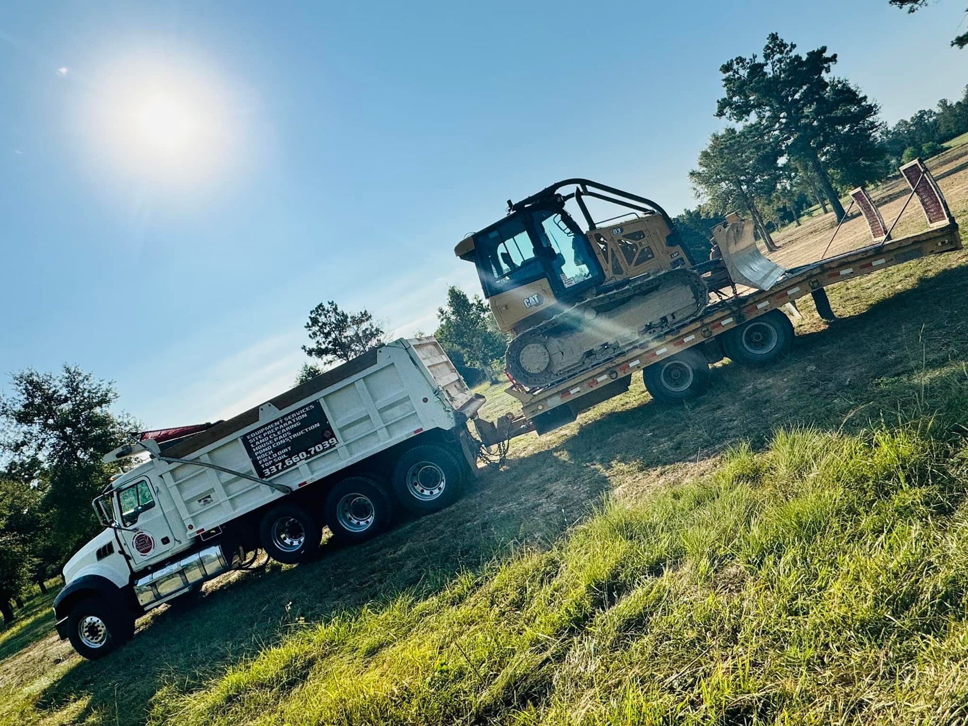 A dump truck and bulldozer on a trailer on a grassy field on a sunny day.