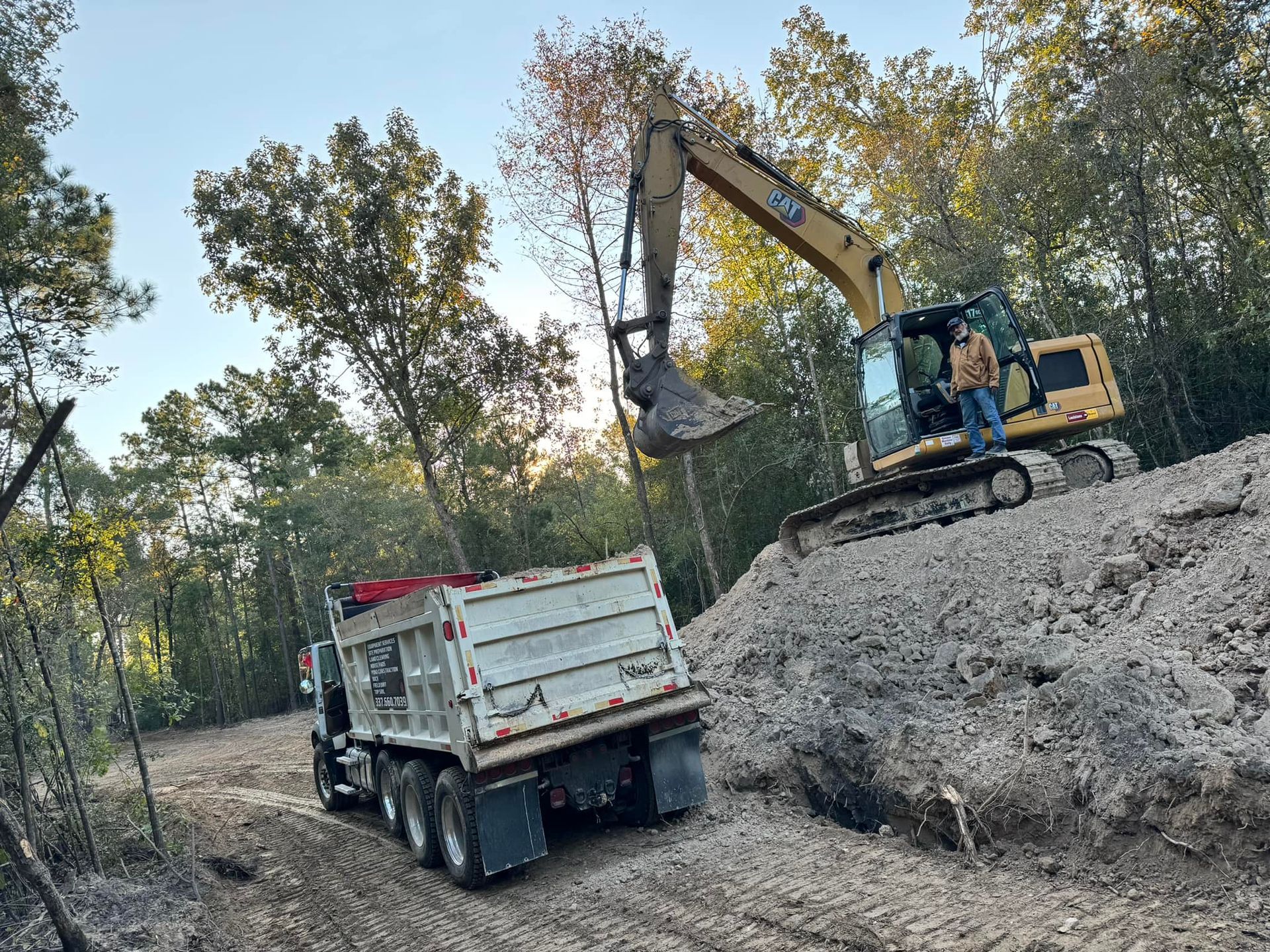 Excavator loading dirt into a dump truck on a dirt road, trees in background.