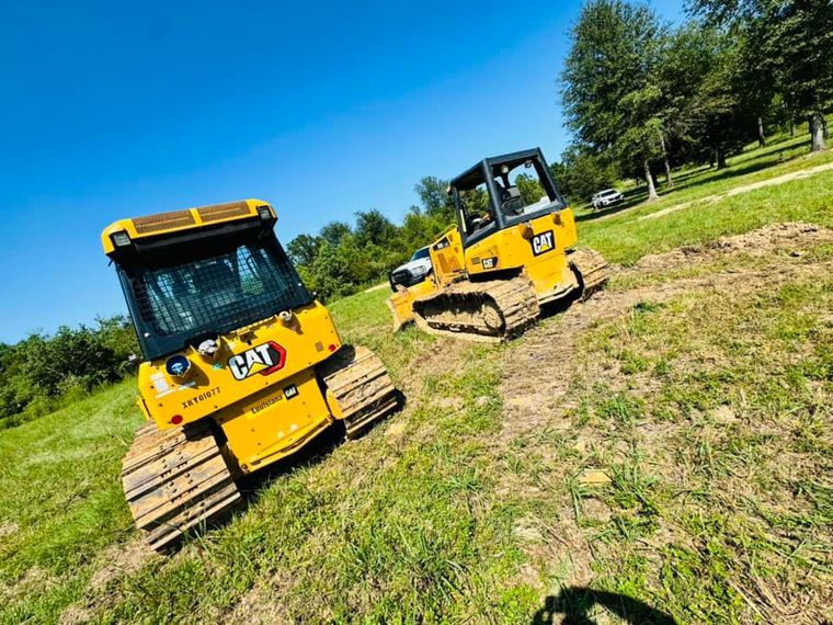 Two yellow Caterpillar compact track loaders on grassy slope under blue sky.