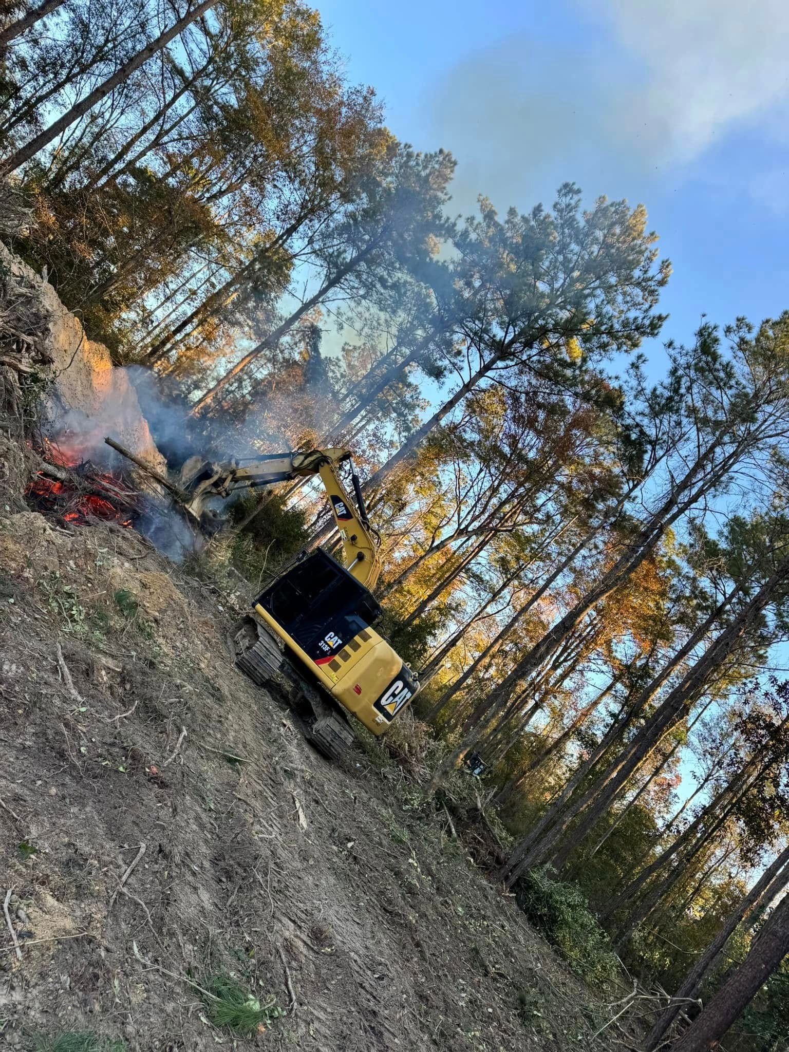 Yellow excavator near burning brush pile in a wooded area with trees and blue sky.