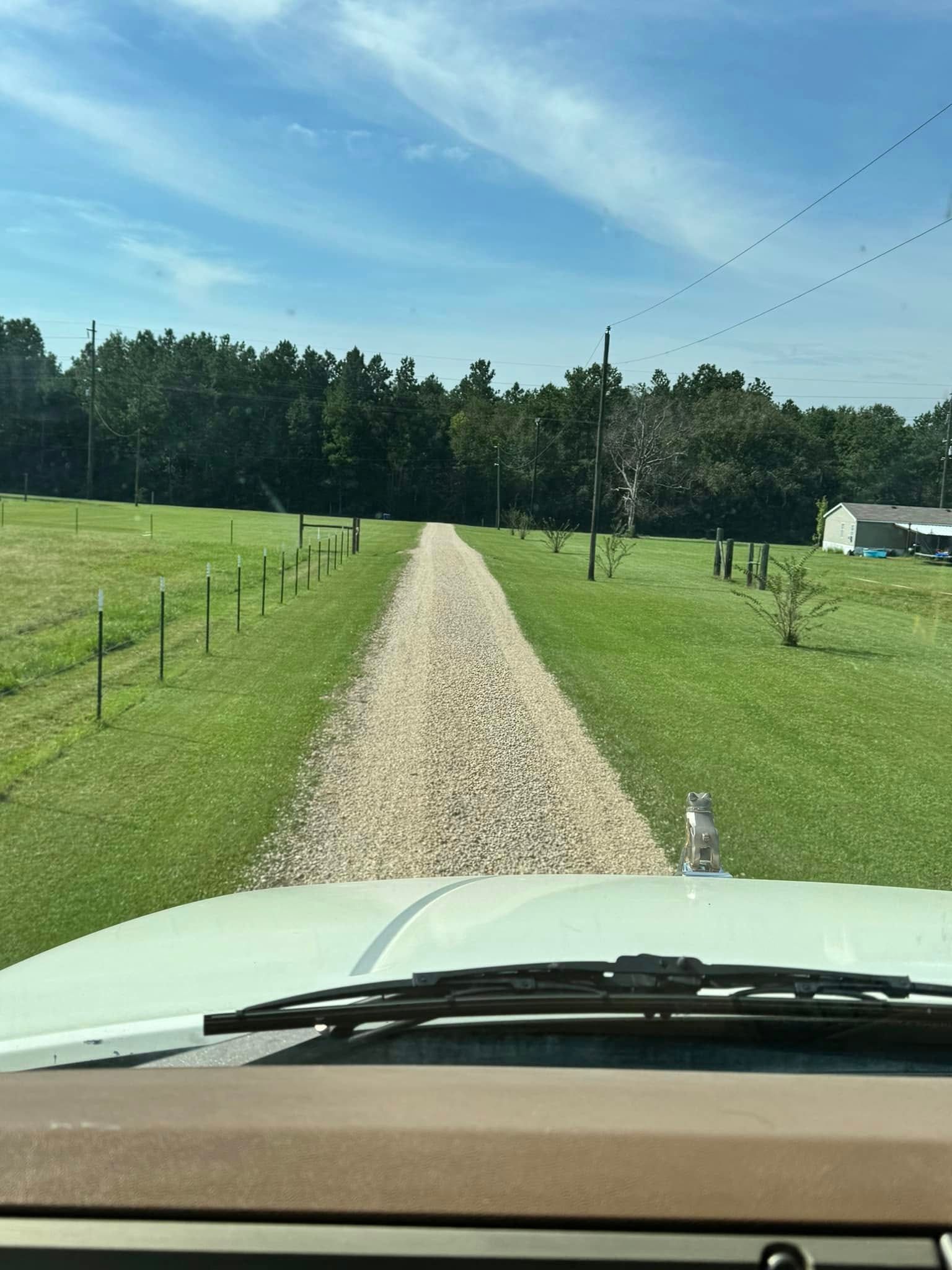 Gravel driveway lined with fence posts, trees, and grass, viewed from inside a vehicle under a blue sky.