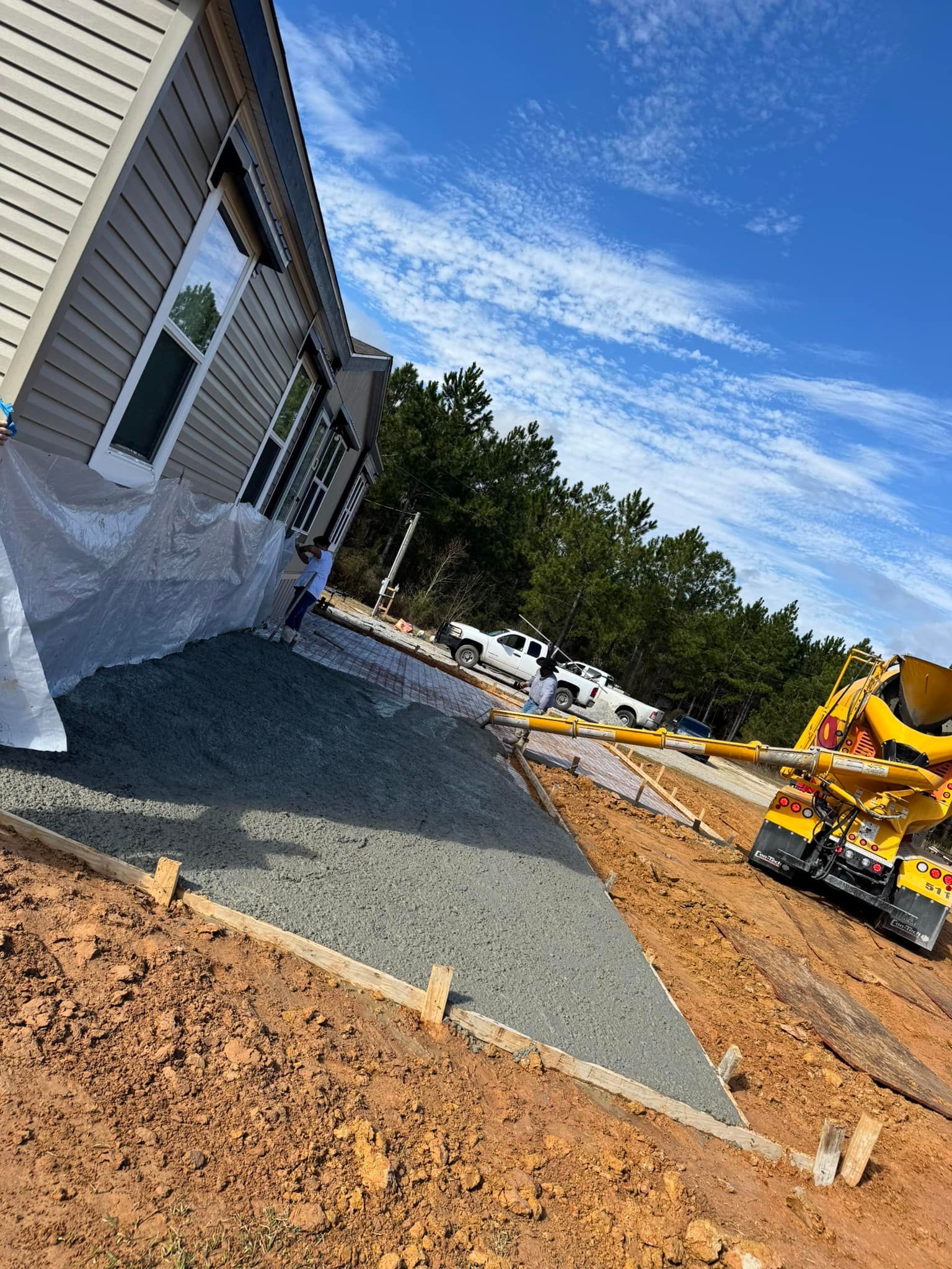 Concrete being poured for a driveway, beside a house under construction; blue sky, dirt, and a cement truck are visible.