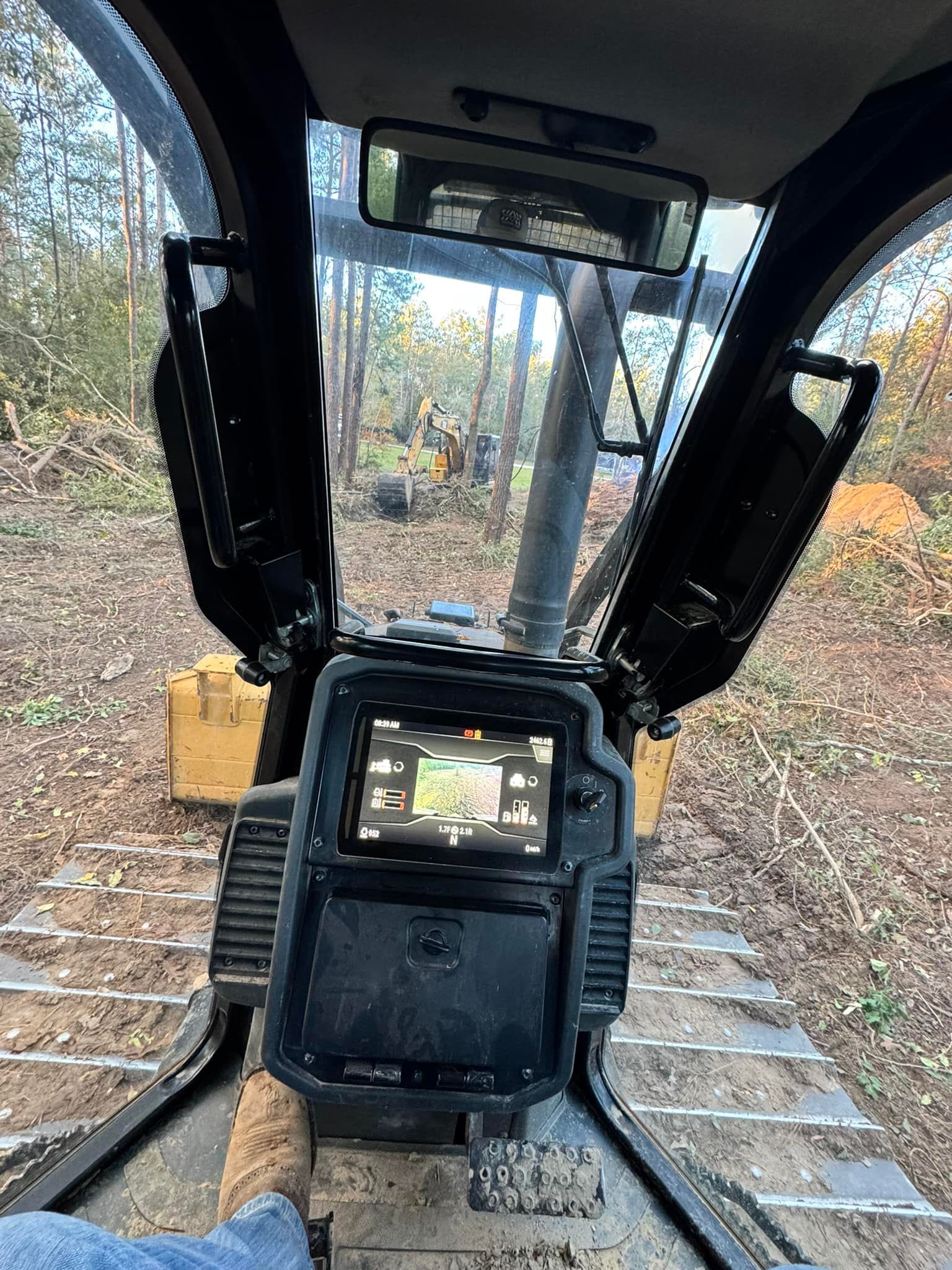 View from inside a heavy machinery cab, looking out at a wooded area.  A screen and controls are in focus.