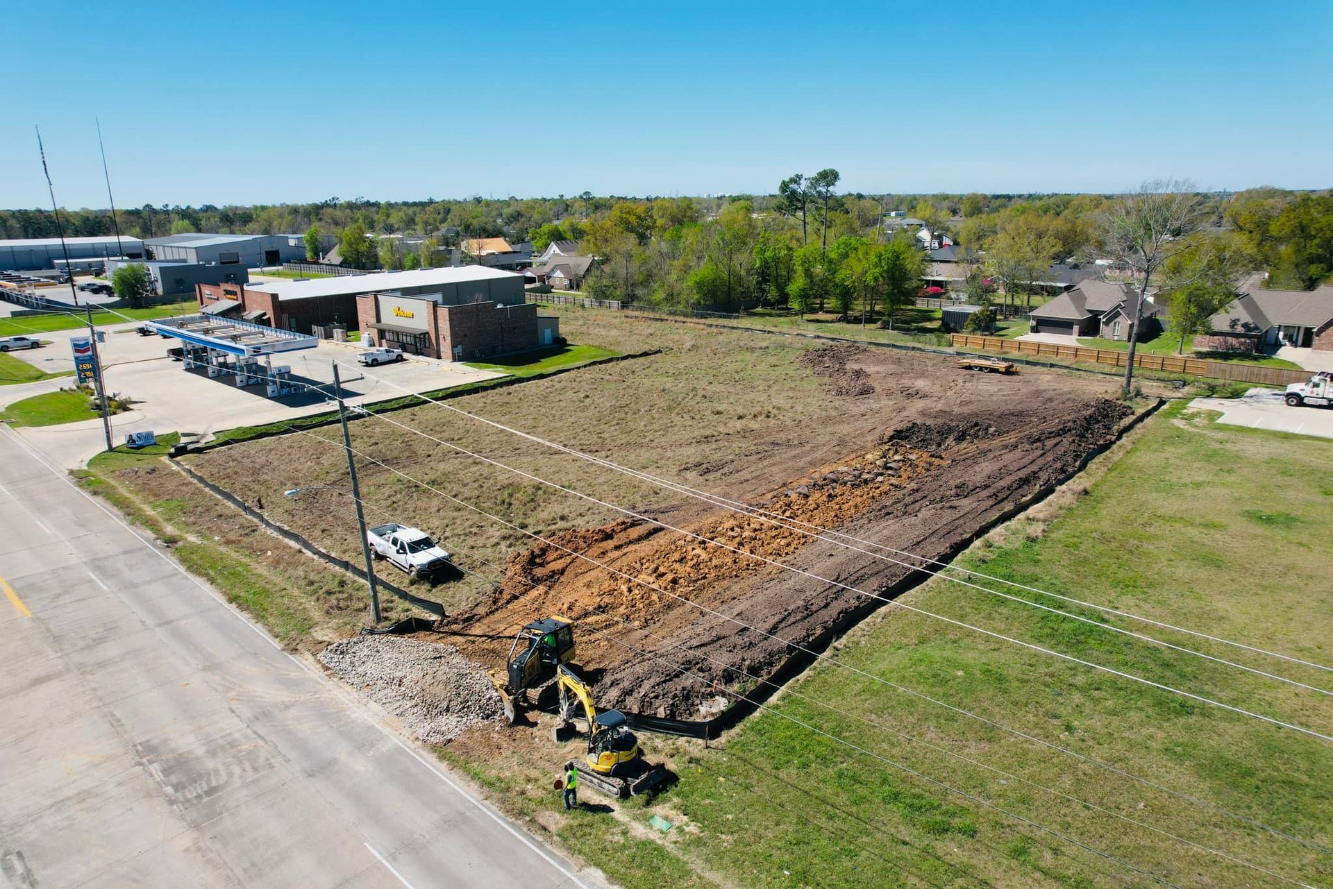 Aerial view of construction site with an excavator, brown soil, and surrounding buildings.
