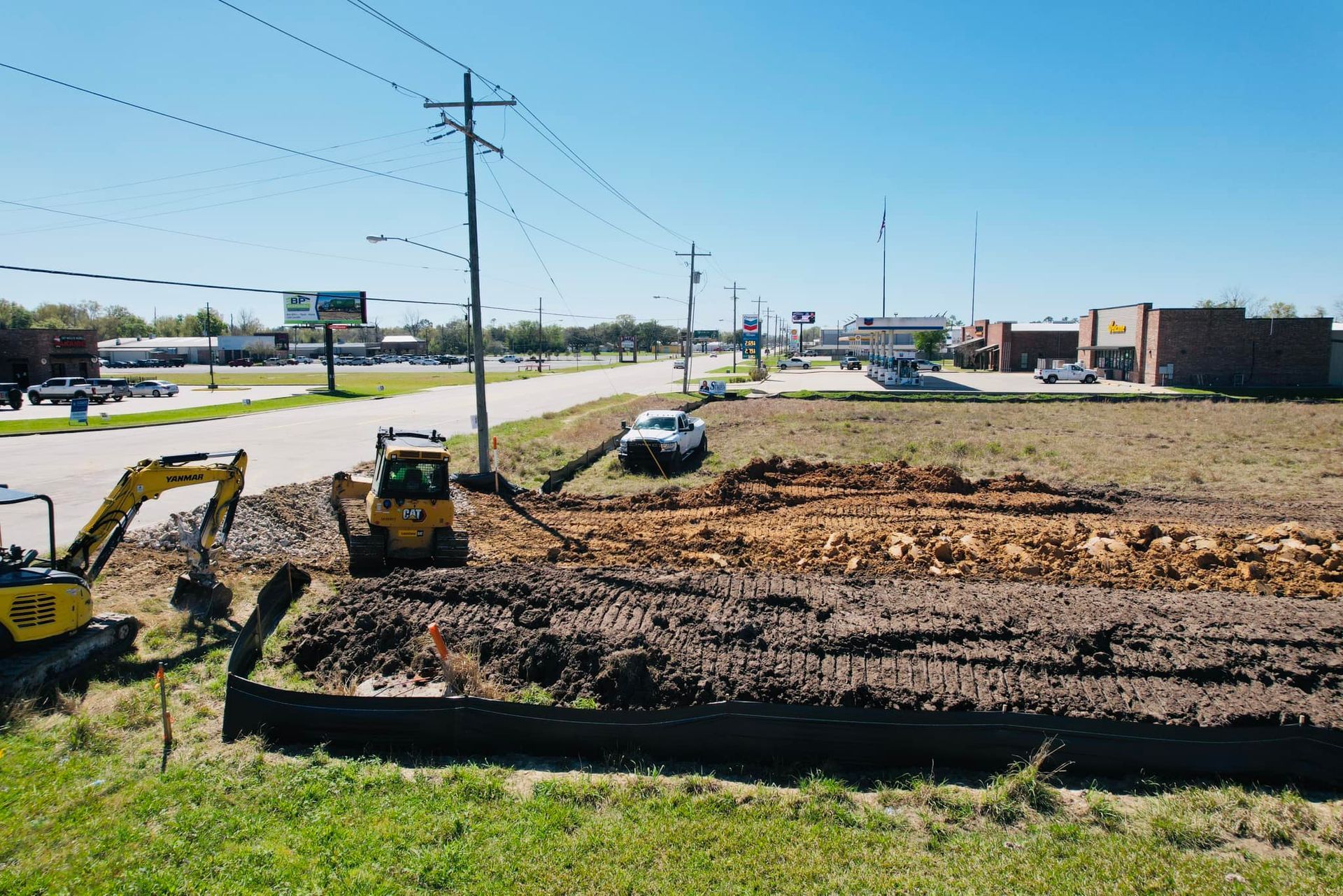 Construction site with excavators and a truck on a cleared, brown dirt lot next to a road and power lines.
