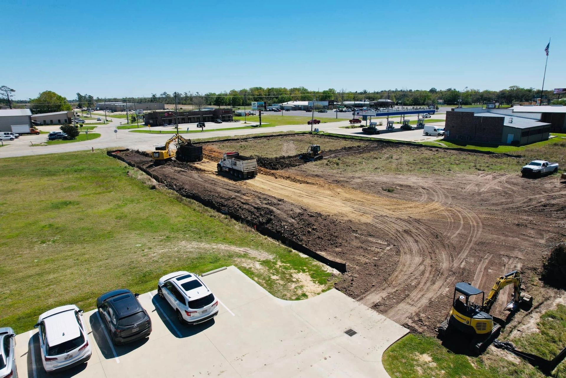 Construction site with earth-moving equipment excavating dirt; cars parked in foreground; buildings and blue sky in background.