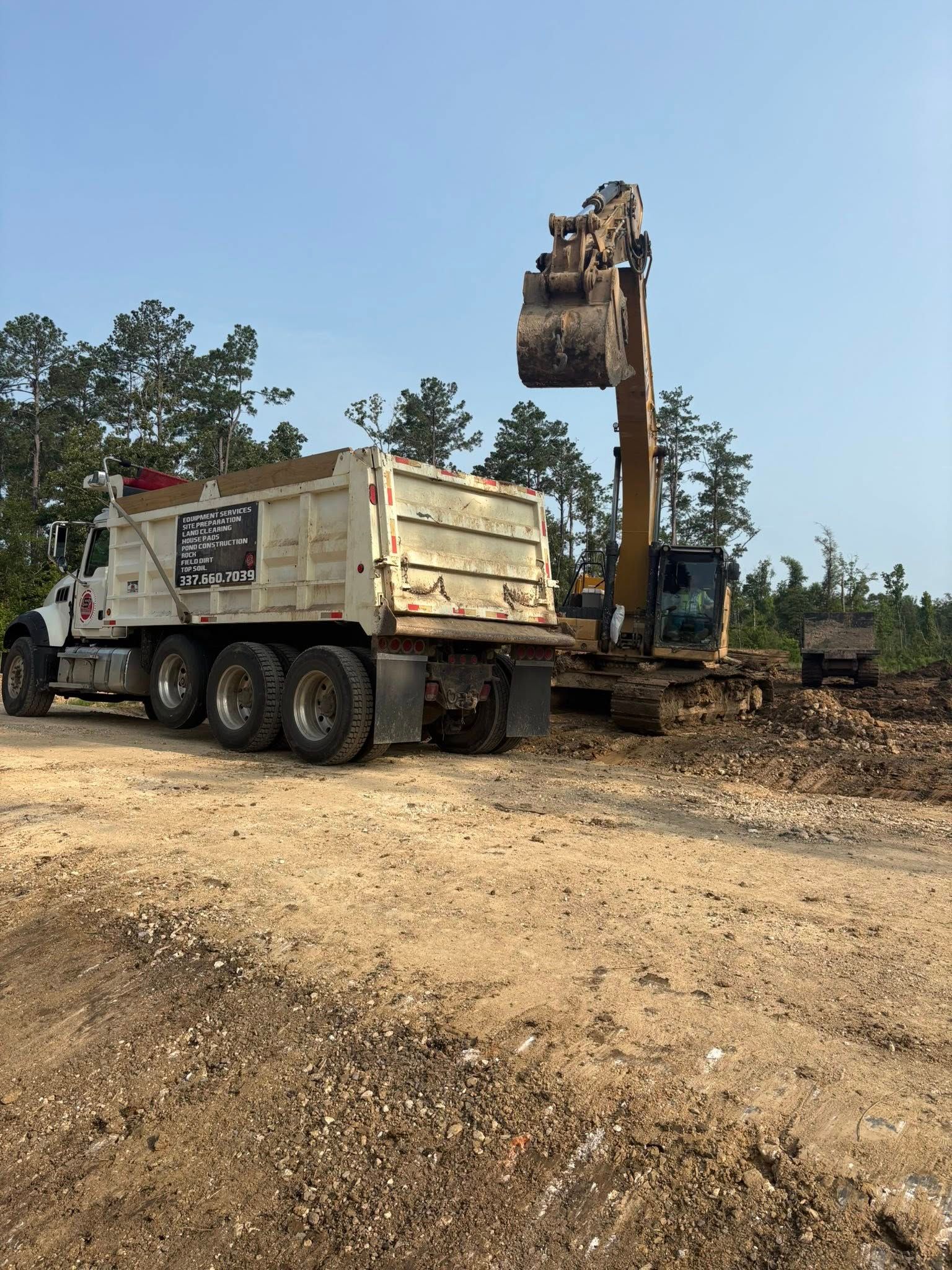 An excavator loading dirt into a dump truck on a construction site. Blue sky and trees in the background.