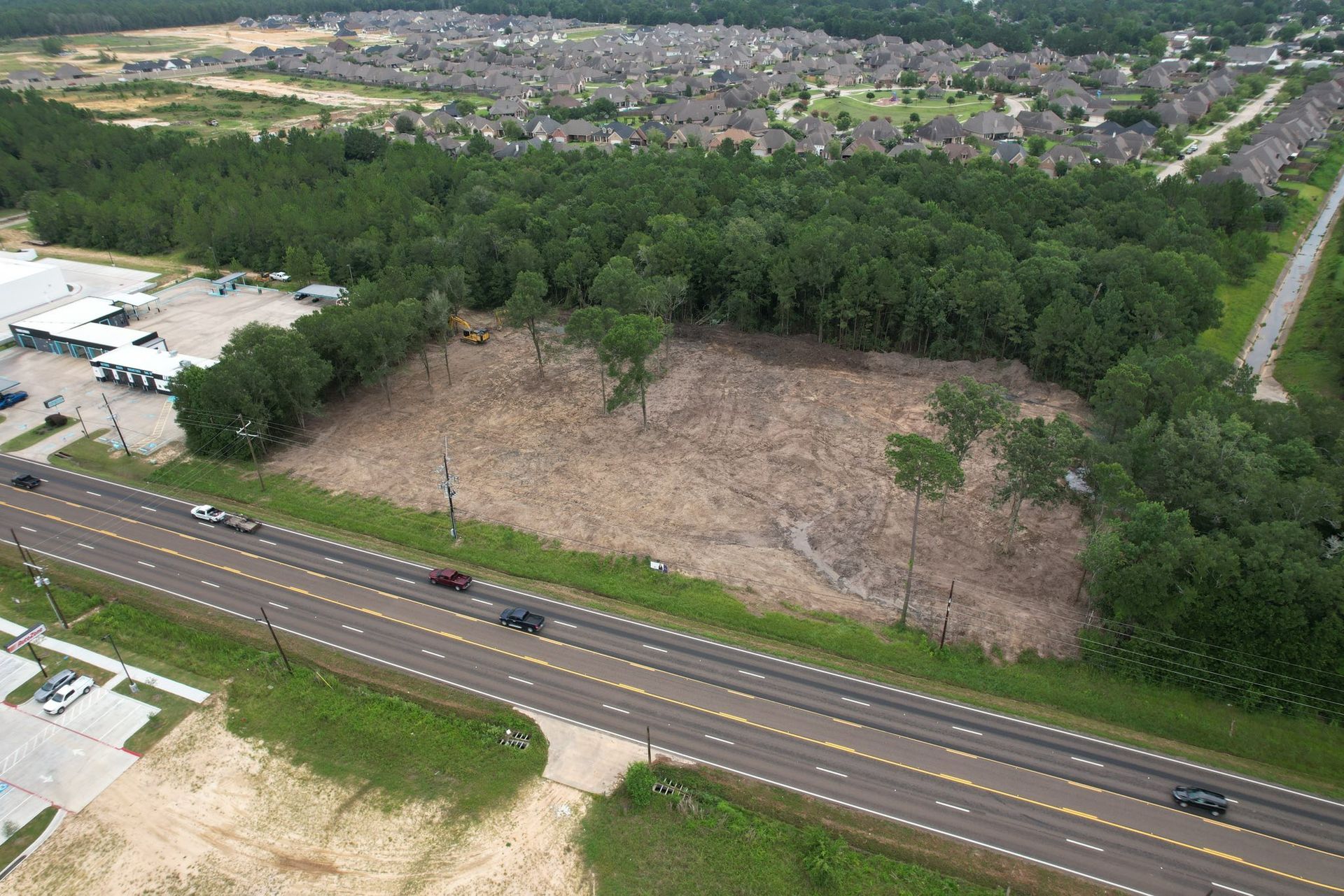 Aerial view: cleared lot next to a highway, surrounded by trees and a residential area.