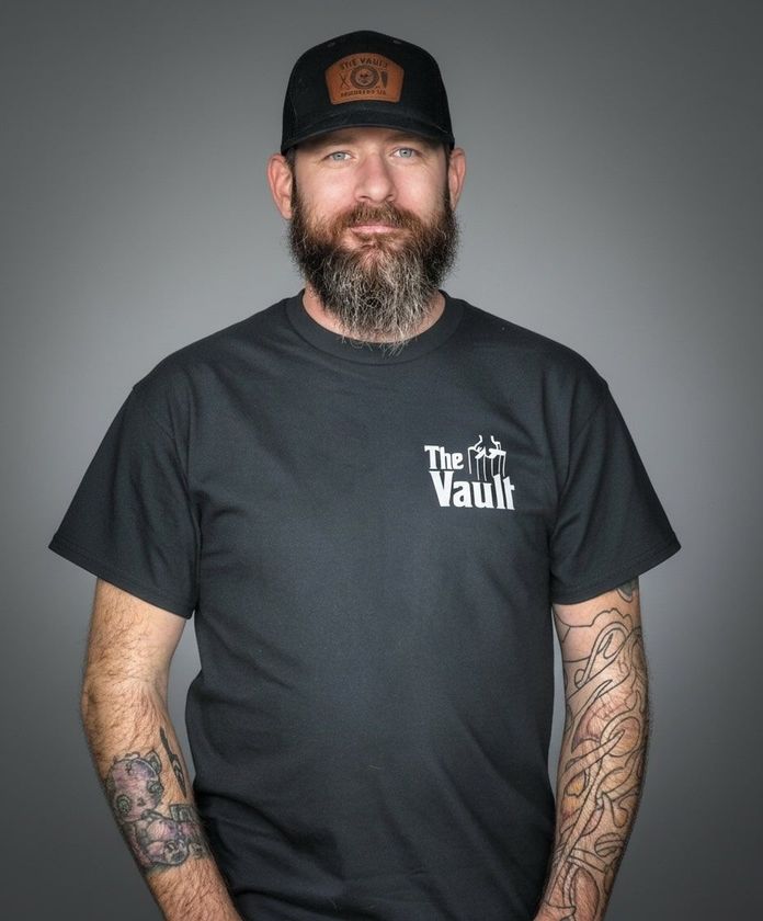 Man with beard and tattoos wearing a hat and t-shirt, stands in front of a gray backdrop.
