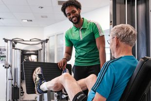Een man helpt een oudere man beenoefeningen te doen in een sportschool.