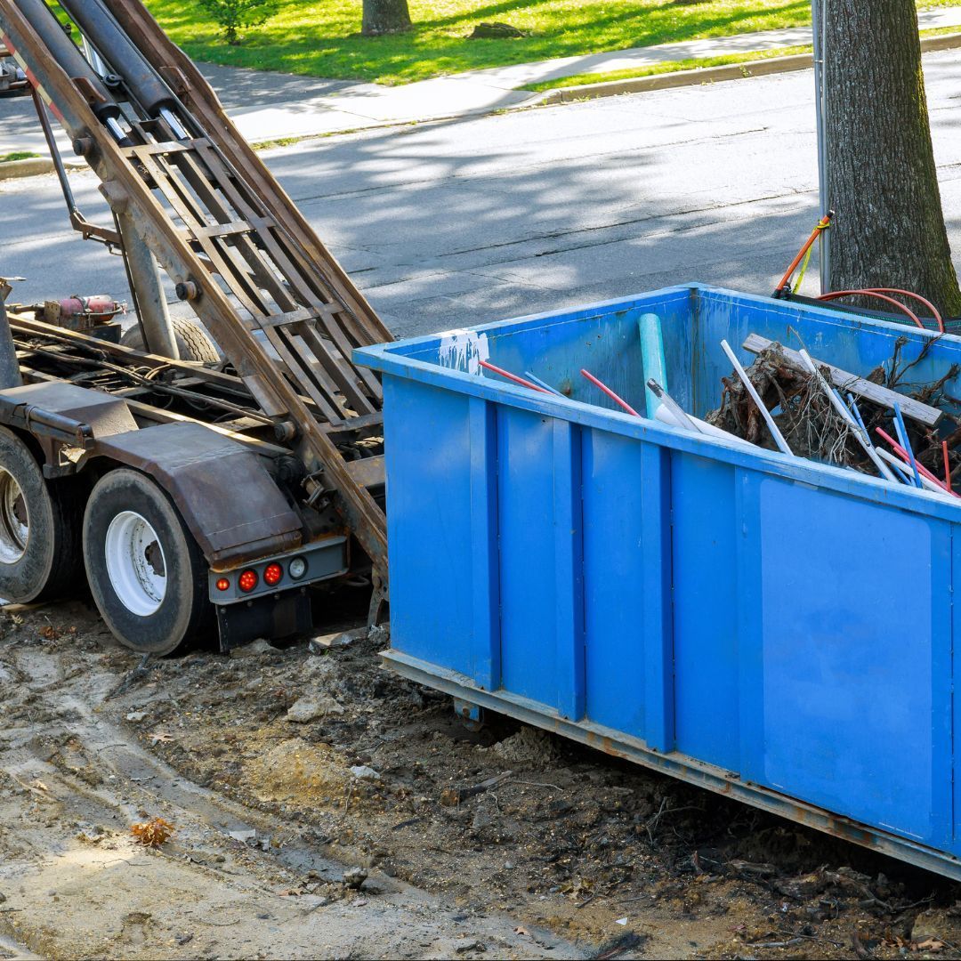 Blue construction dumpster beside a trailer-mounted ladder on a roadside work site