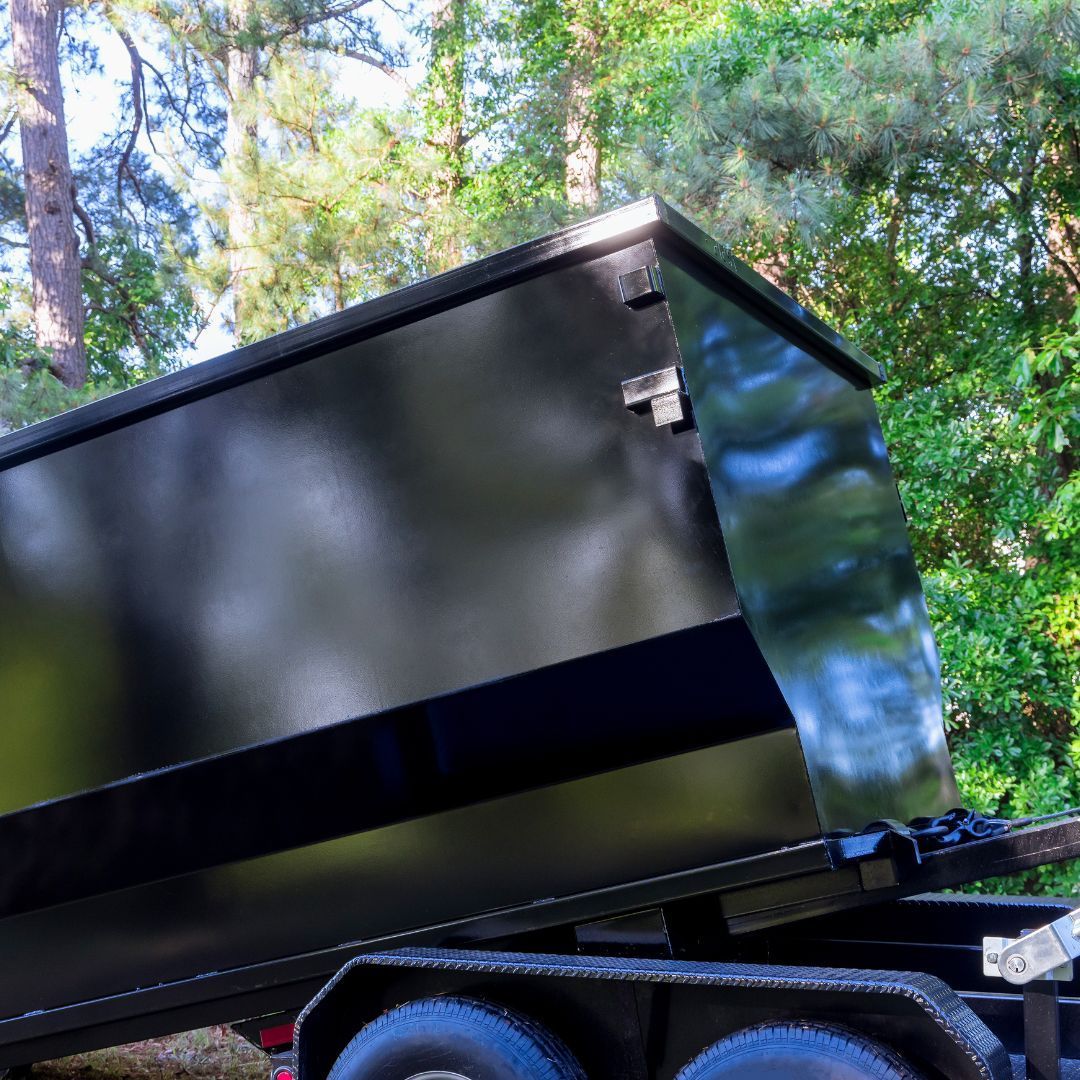 Black dump trailer parked outdoors in a wooded area, with a blue trim and raised side panels