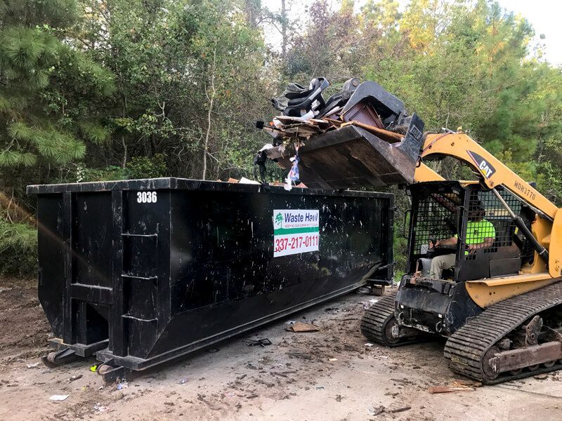 Yellow skid steer loading debris into a black dumpster in a wooded area.