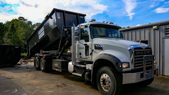 White Mack dump truck hauling a black dumpster trailer in an outdoor industrial yard