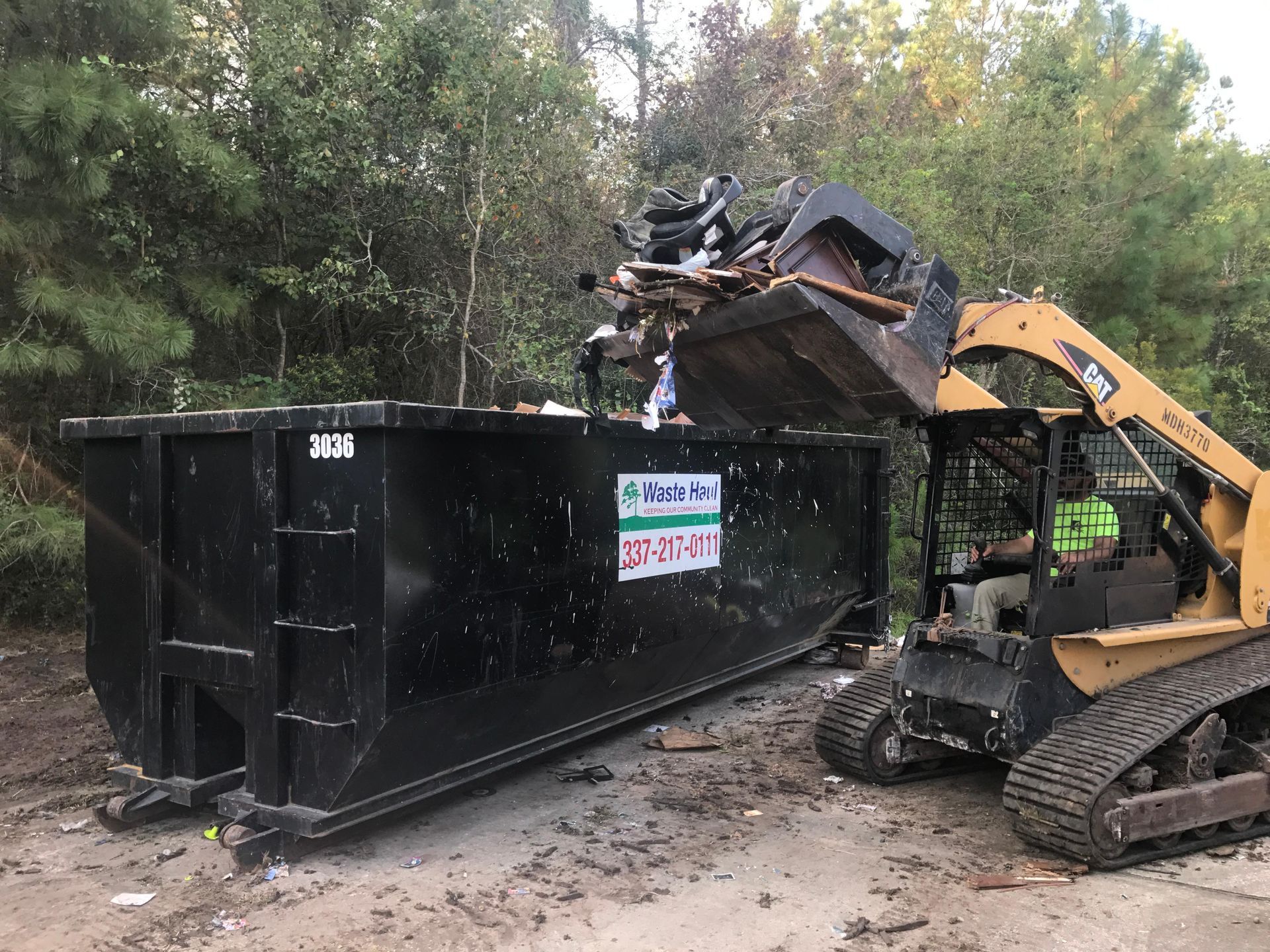 Yellow excavator loading debris into a large black dumpster in a wooded outdoor area
