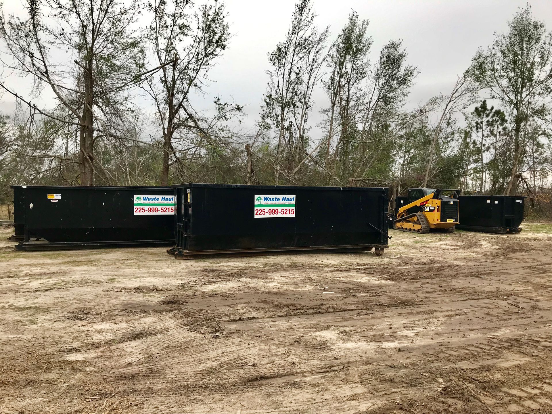 Black dumpsters and a yellow skid steer on a dirt lot with leafless trees in the background