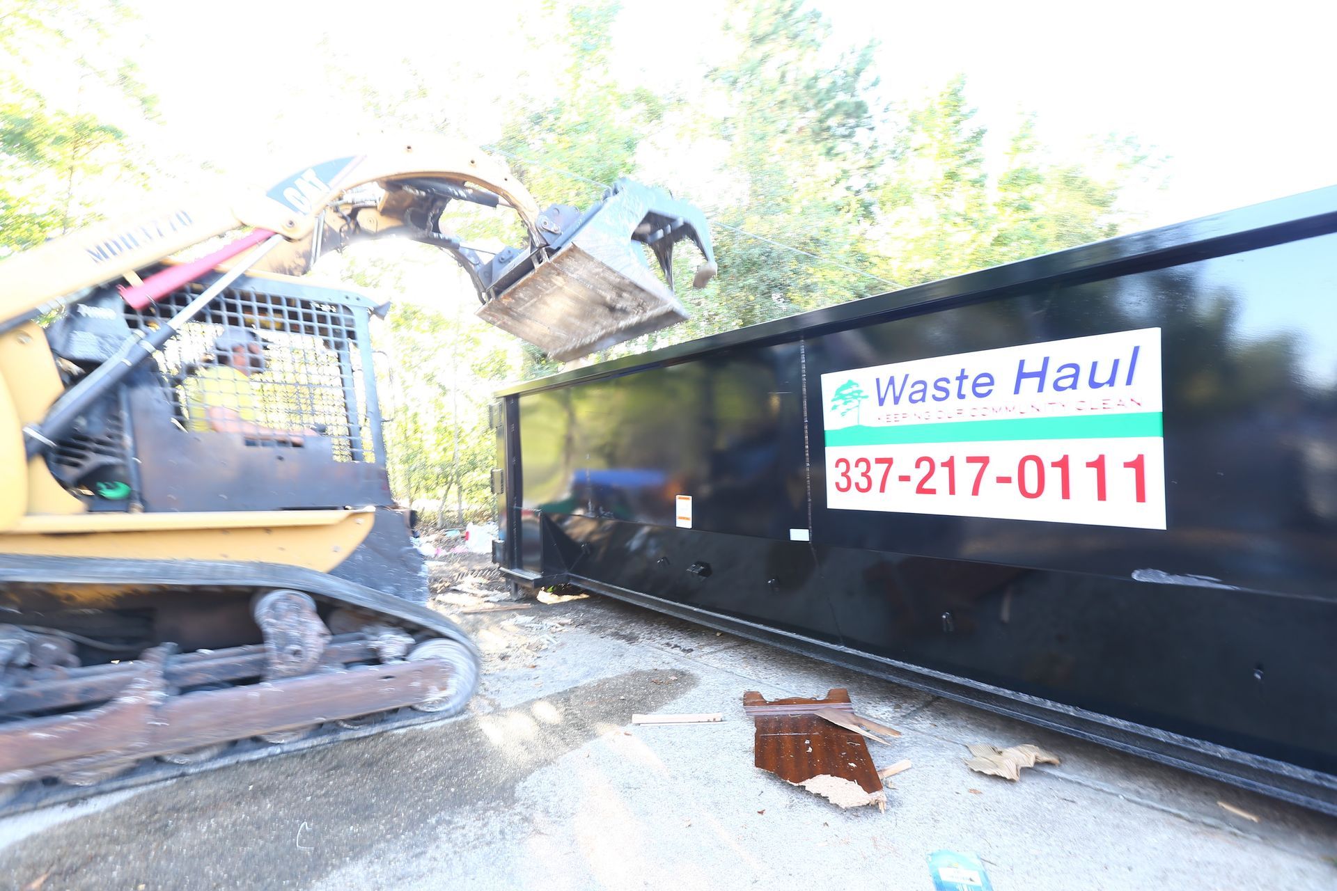 Excavator loading debris into a black waste haul dumpster with a visible phone number sign.