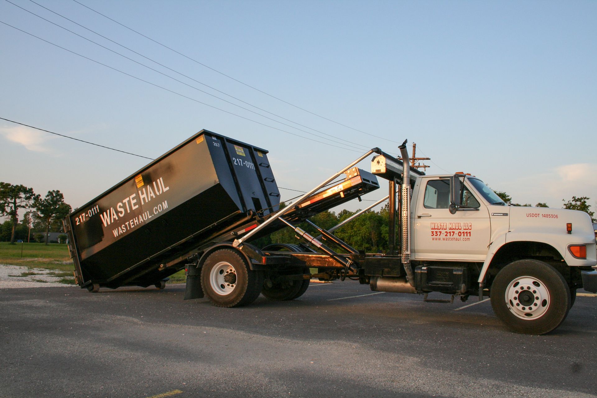 White dump truck tipping a black container on a roadside at dusk