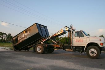 Dump truck tilting a black dumpster bin on a road at dusk